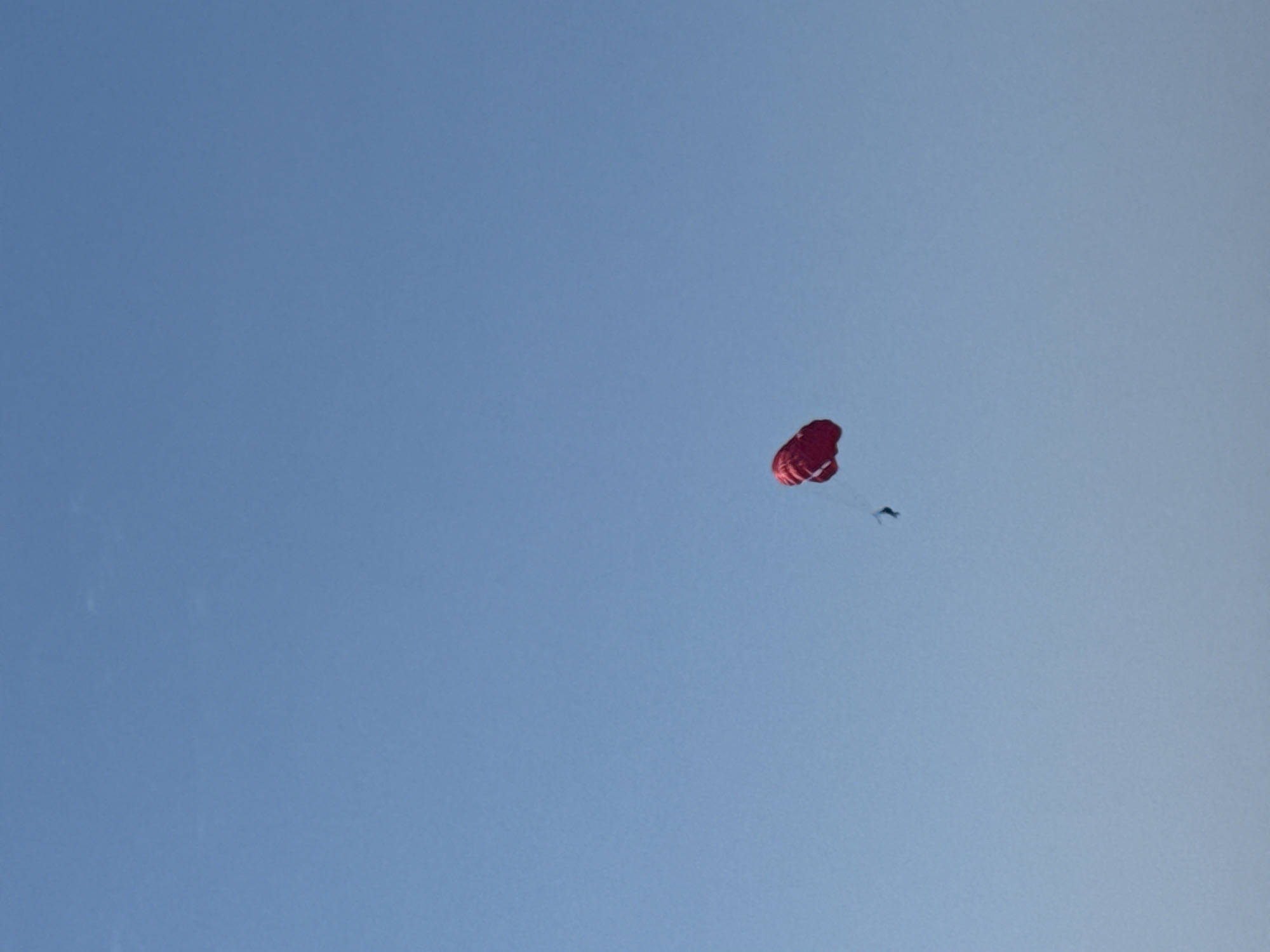 A bright red parasail with a small dark figure underneath is suspended against a clear, pale blue sky. The parasail appears inflated and is gently drifting. The scene suggests a daytime activity, likely recreational, given the clear weather. The location is identified as Boreti, Montenegro, which is a coastal area, implying the parasail activity might be taking place over or near the Adriatic Sea. There is no visible text in the image. The overall mood is serene and peaceful, with the vast expanse of the sky dominating the frame.