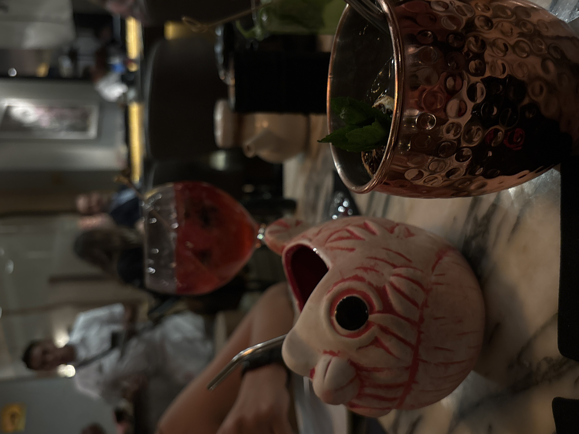 The image, taken in Mexico City, Mexico, displays a close-up of two cocktails on a marble-patterned table in a dimly lit bar or restaurant environment, rotated ninety degrees clockwise. In the immediate foreground, a distinctive white ceramic mug shaped like a fish is featured, adorned with red painted patterns resembling scales or tribal markings, and prominent black eyes. A silver-colored metal straw extends from the fish's mouth. A person's arm and hand are partially visible, holding the base of this unique mug.
To the upper left, a hammered copper mug, commonly used for a Moscow Mule, contains a green liquid, fresh mint leaves, and a lime wedge, indicating another cocktail. Both drinks are in sharp focus, contrasting with the blurred background. The background reveals indistinct figures of people, likely patrons, and warm, soft lighting, suggesting an evening or nighttime setting. The scene captures a social occasion where individuals are enjoying creatively presented beverages. No text is visible in the image.