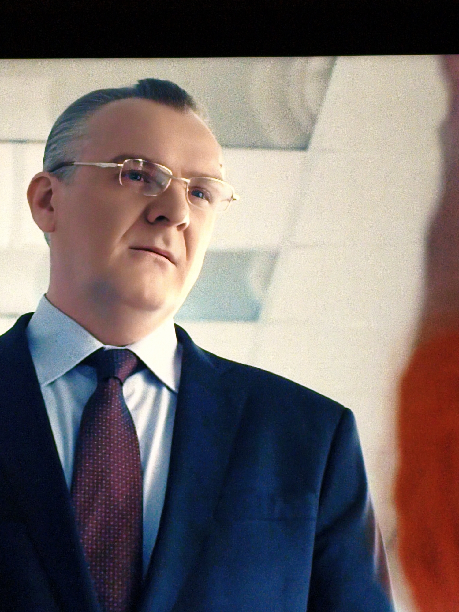 A man wearing a dark blue suit, white dress shirt, and a burgundy patterned tie is shown in medium close-up, looking slightly to his left. He is wearing gold-rimmed glasses and his hair is slicked back. The background appears to be an indoor environment with a light-colored wall and ceiling, possibly an office or a well-lit hallway. A blurry orange object is visible to the right of the frame. There are no discernible landmarks or elements that would indicate a specific location within Vladivostok, Russia.