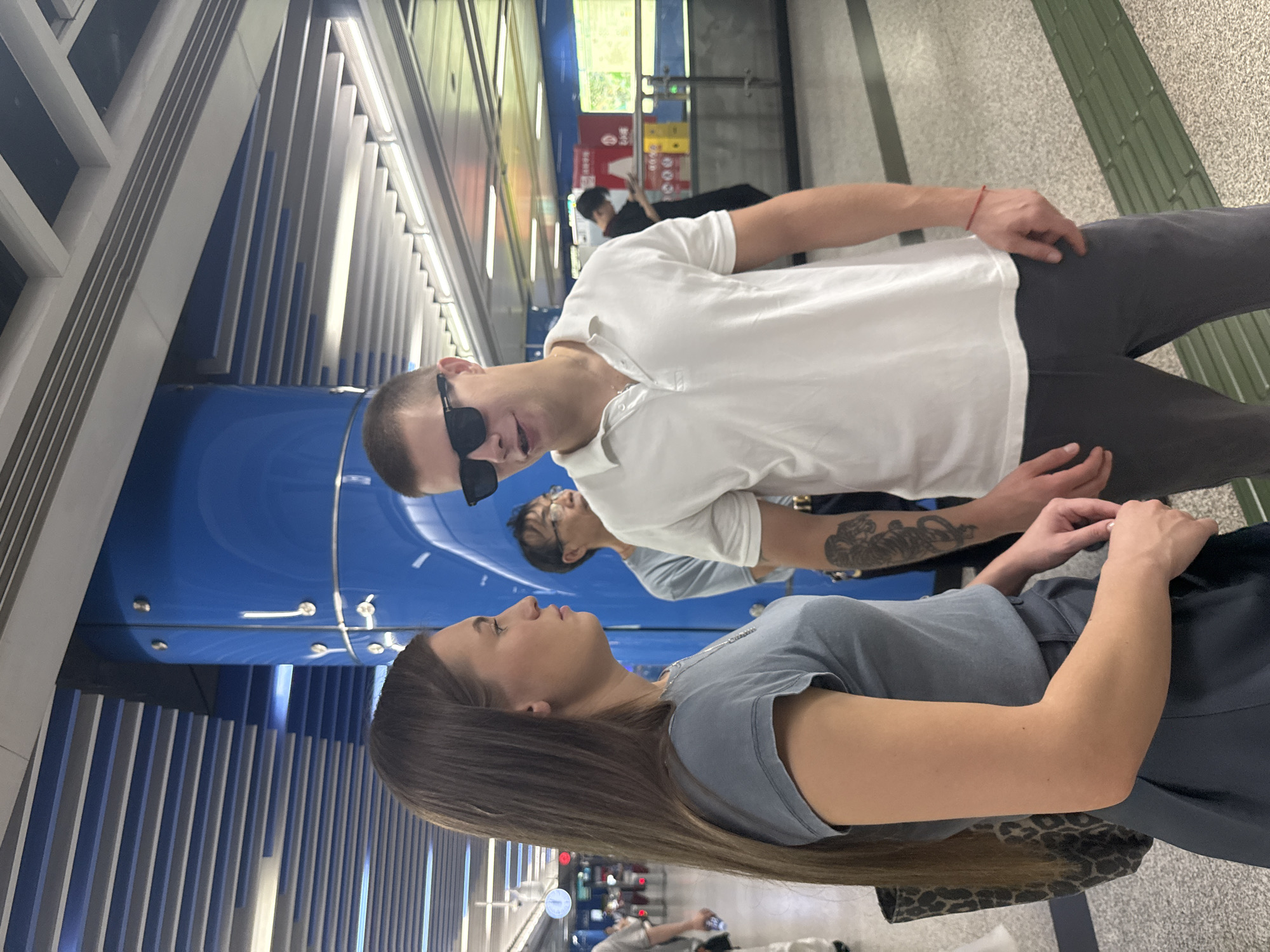 A medium shot shows a woman and a man standing in what appears to be a subway station in Haizhu District, China. The woman, on the left, has long, brown hair and is wearing a grey t-shirt and a dark blue skirt. She is carrying a leopard-print bag. The man, on the right, is wearing a white polo shirt, sunglasses, and dark grey pants. He has a red string bracelet on his wrist and a tiger tattoo on his left arm. They are facing each other and appear to be in conversation. In the background, other people are visible, along with the station's blue columns and ceiling.