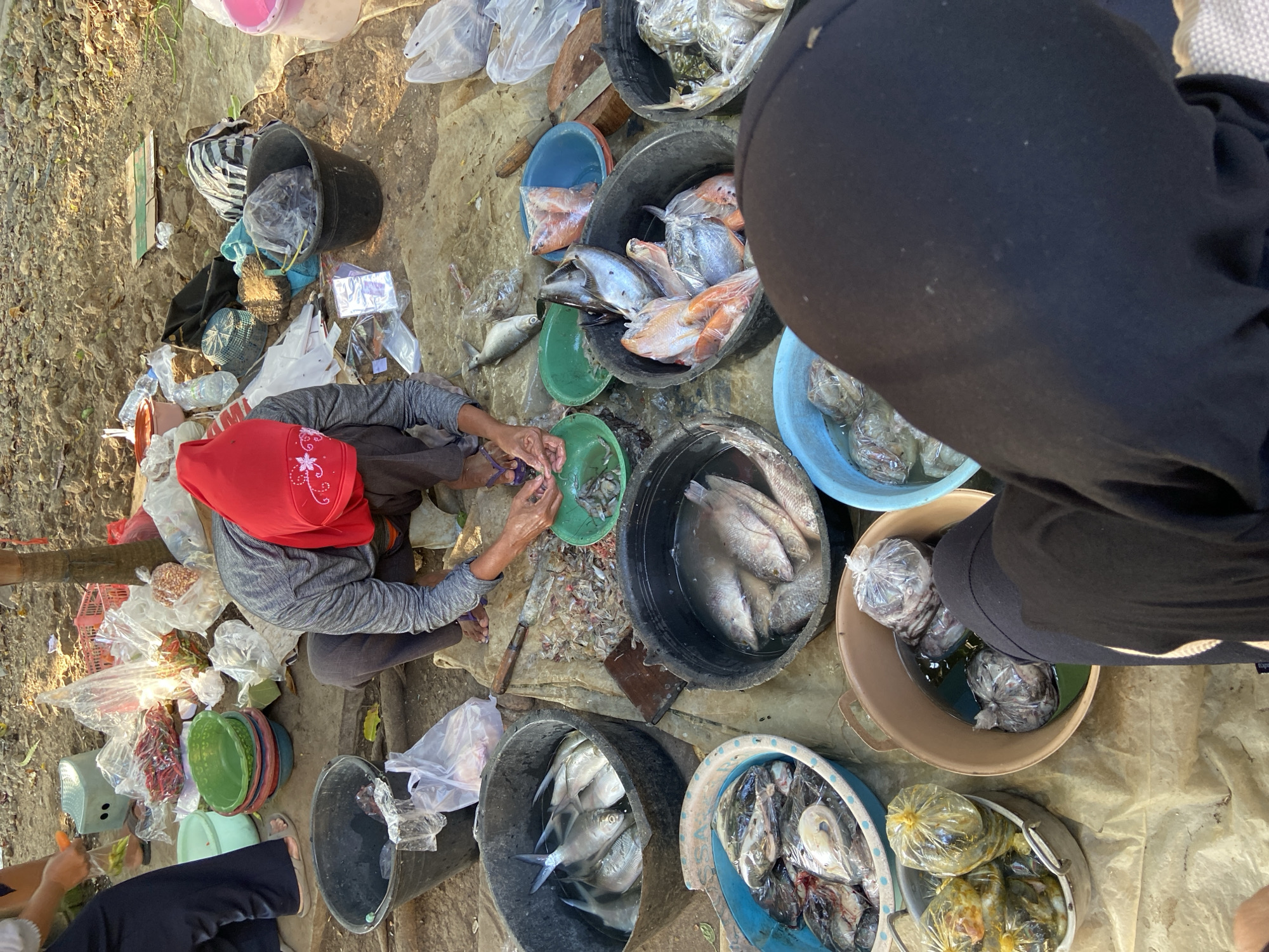 A street market scene is captured in Kecamatan Pati, Indonesia. A woman, wearing a red headscarf and grey long-sleeved shirt, kneels on the ground, processing seafood. She is using her hands, with a small green bowl in front of her containing what appears to be shrimp or small fish. Surrounding her are various containers filled with fish and other seafood. Black buckets hold large quantities of fish, some with visible scales and fins. A blue plastic basin contains more fish, some wrapped in plastic. Another beige basin holds several plastic-wrapped packages of seafood. In the background, red plastic crates and bags of produce like chilies are visible. The ground is a mix of dirt and what appears to be a tarp or cloth covering. The overall environment suggests an outdoor market setting.
