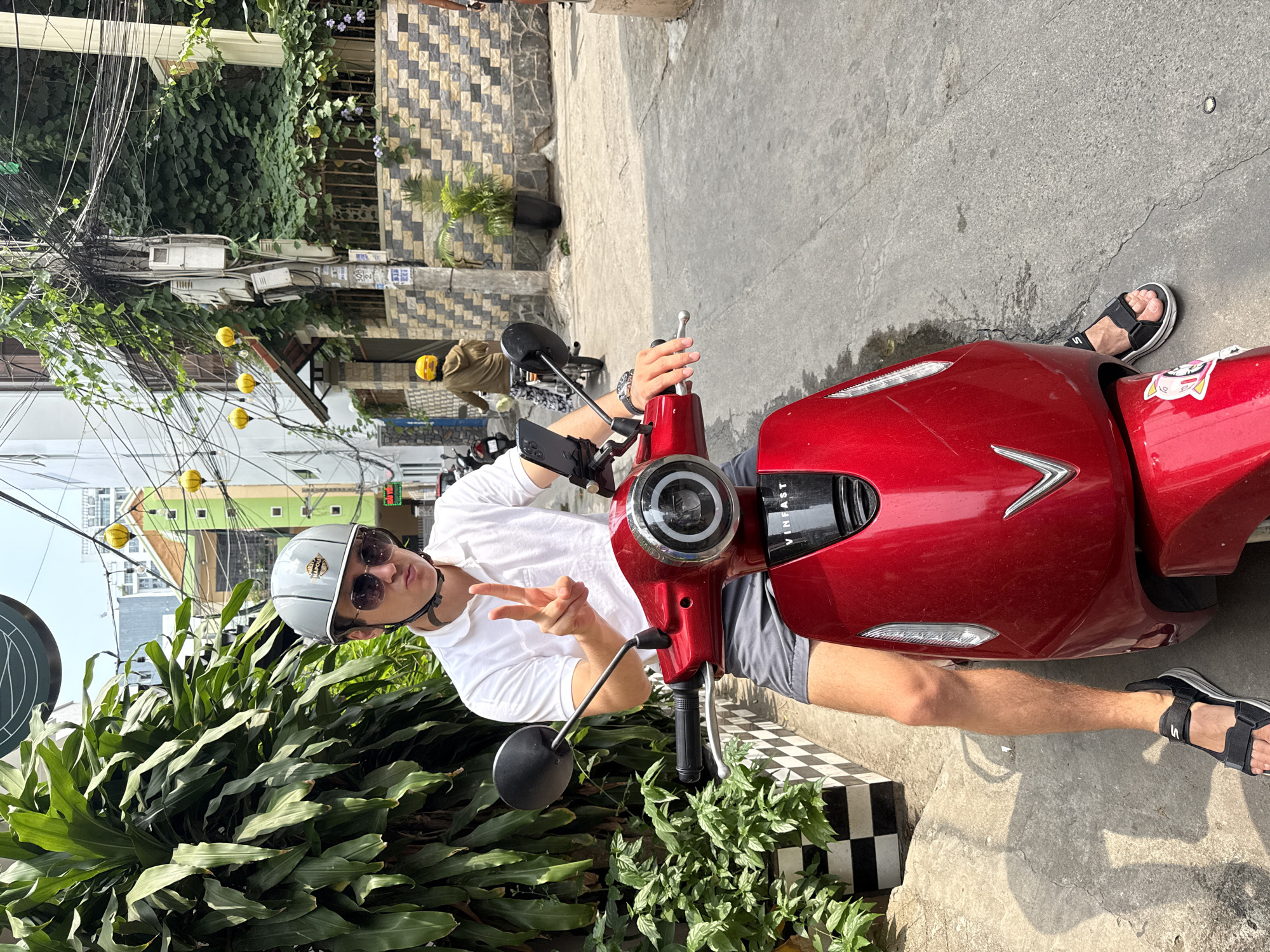 A young adult male, wearing a white t-shirt, gray shorts, a light gray helmet, and sunglasses, is posing on a red VinFast electric scooter on a city street. The location is Nha Trang, Vietnam, judging by the architecture and signage in the background. He is giving a peace sign with his left hand. He is wearing black sandals. The scooter is parked next to a planter with green foliage. Behind him are buildings, power lines, and other parked scooters. The street is paved with gray concrete. A house is visible with a geometrically patterned wall.