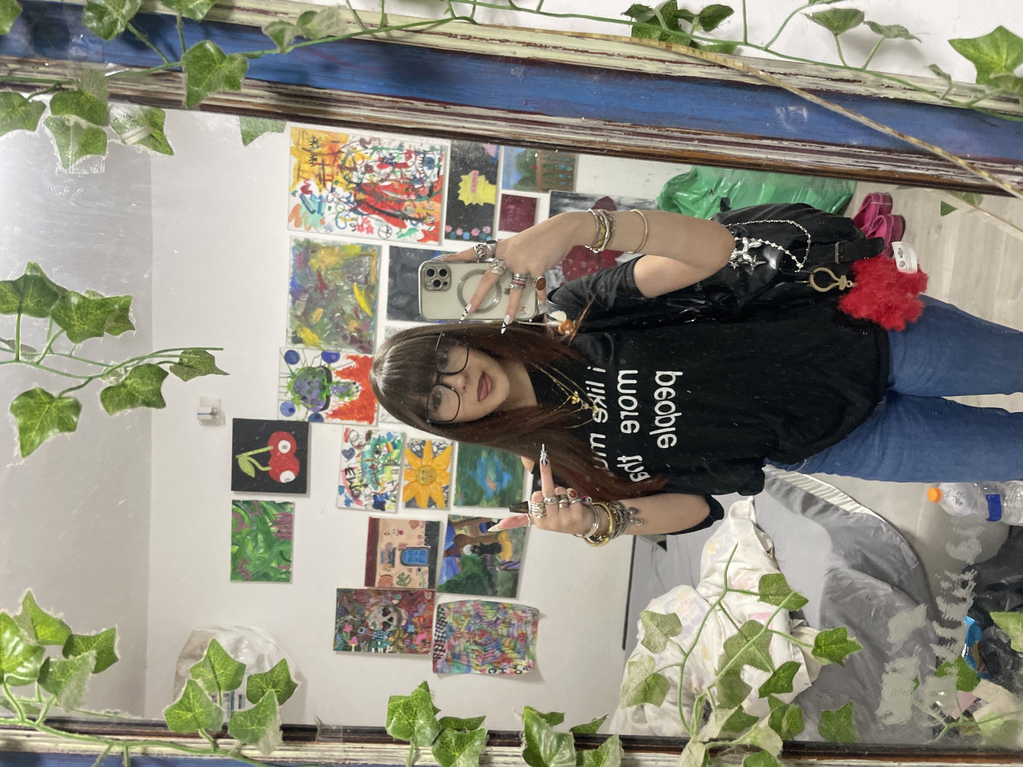 A woman in a black t-shirt and jeans is taking a selfie in a mirror. The mirror has a blue frame and is decorated with artificial ivy. The woman is wearing glasses and has long brown hair. She is also wearing several rings and bracelets. In the background, there are several small paintings on the wall. There is a bed covered with a gray sheet. The location appears to be indoors. The city and country are Raanana, Israel.