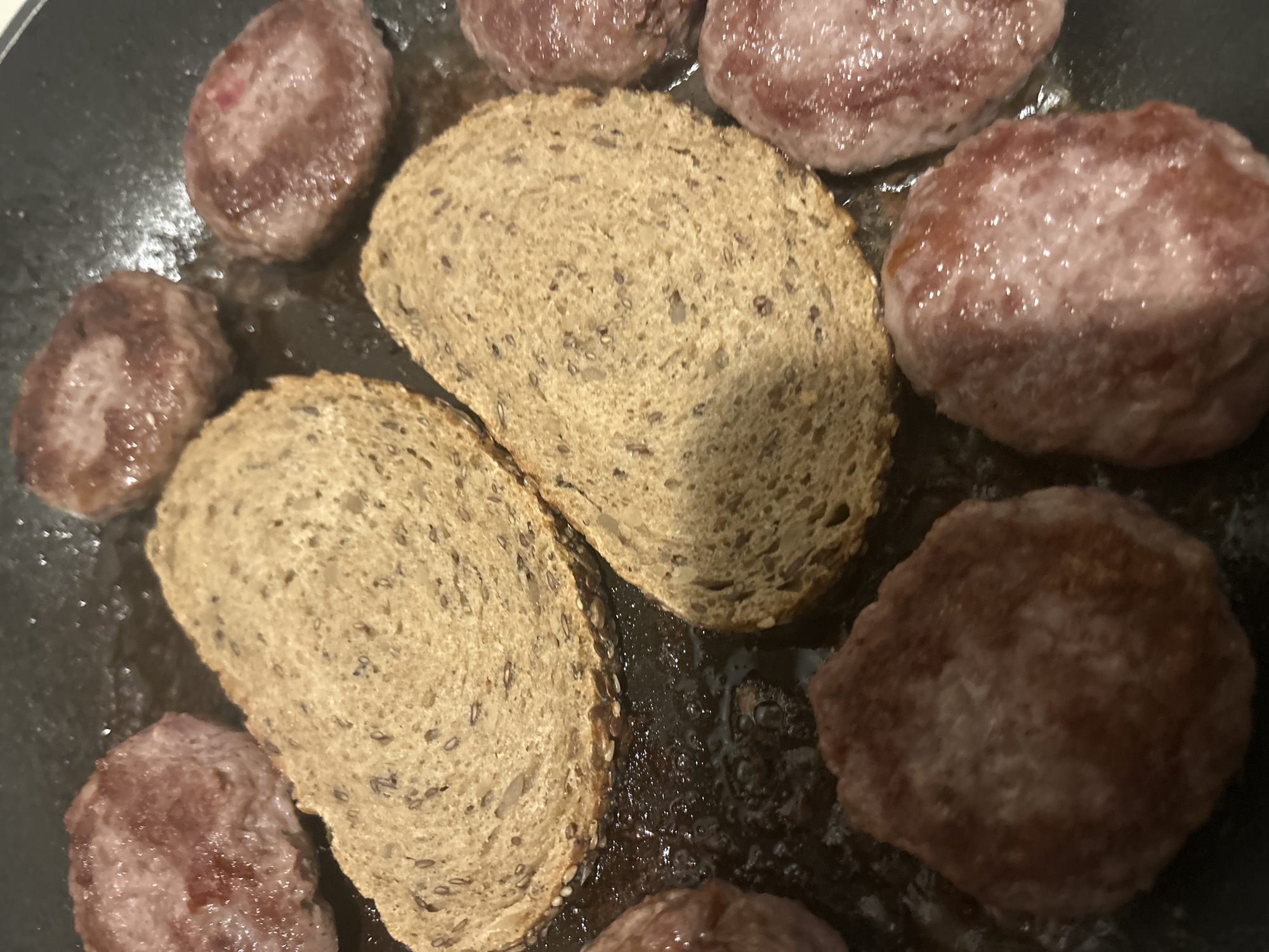 The close-up image captures a cooking scene within a dark-colored pan, located in Varna, Bulgaria. Several small, roundish, reddish-brown items, resembling meatballs or small burger patties, are sizzling in rendered fat or oil. Adjacent to these meat items are two slices of light brown, seedy bread, likely multi-grain, also cooking in the pan and absorbing some of the juices. The composition focuses intimately on the food preparation. There are no people or visible text, and the time of day or external weather conditions cannot be determined from the indoor setting.