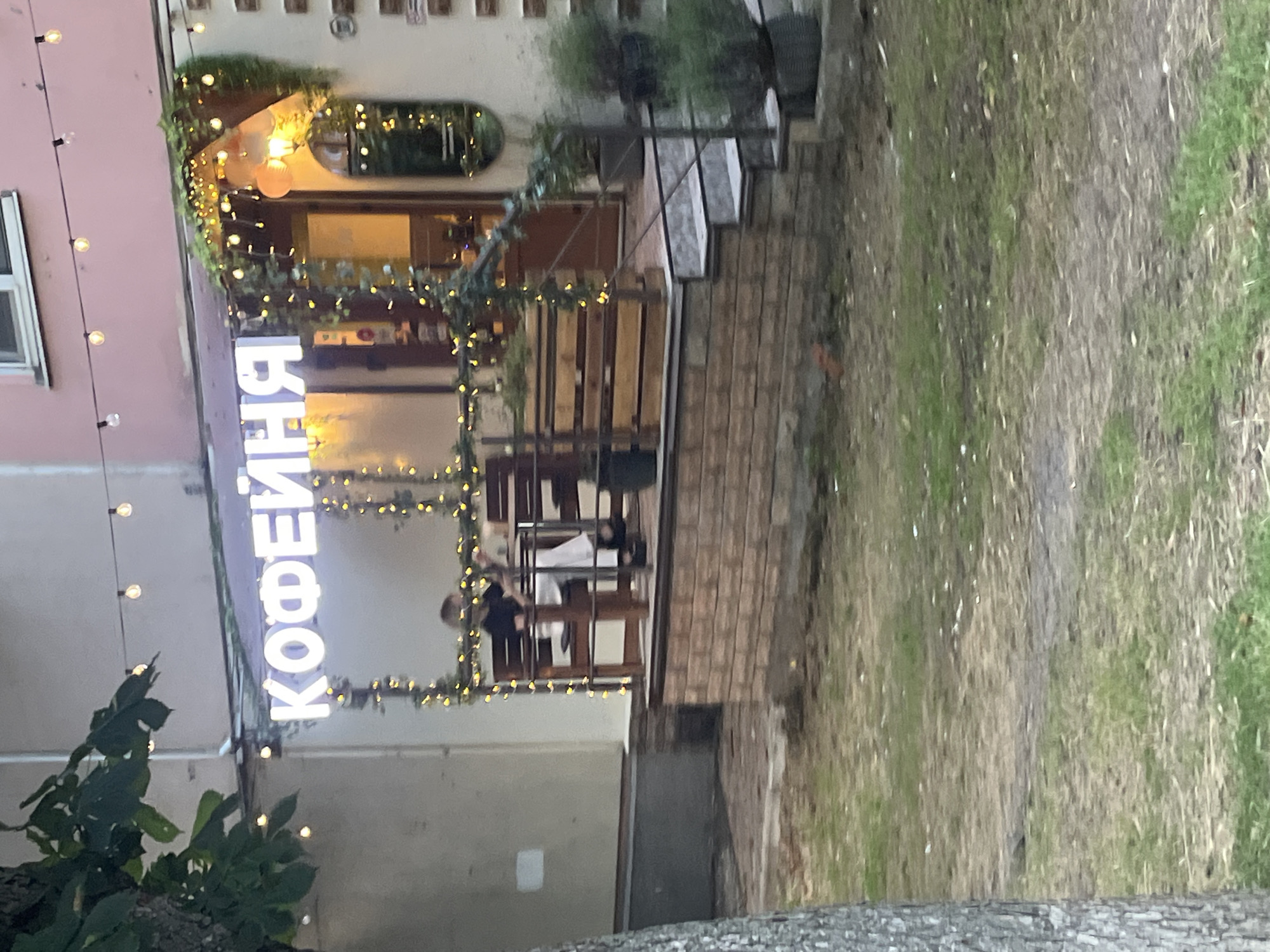 The image shows the exterior of a cafe with a large illuminated sign that reads "КОФЕЙНЯ" in Cyrillic letters. The cafe has an outdoor seating area with wooden tables and chairs, decorated with string lights and greenery. A person is seated at one of the tables. The cafe is located in the city of Krasnodar, Russia. The scene is captured in daylight, and the overall environment appears to be an urban setting with a slightly unkempt grassy area in the foreground.