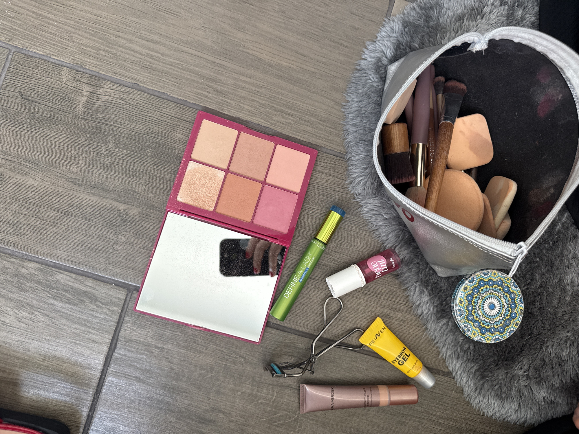 An overhead shot displays an array of makeup and beauty products laid out on a grey wooden floor, suggesting an indoor setting. To the left, a vibrant pink makeup palette is open, revealing six square pans of various shades including light nude, peach, shimmer, and rosy pink, with some powder residue indicating use. Its mirror reflects a hand with dark-painted fingernails.
Adjacent to the palette are several individual products: a green tube of "DEFINE-A-LASH WATERPROOF MASCARA" (brand partially visible as "YBELLI INE", likely Maybelline), a small red-capped bottle of "bene tint" by "benefit", and a silver eyelash curler. Below these, a yellow tube of "PEIVEN EYEBROW GEL" (NET 1 fl oz (30 mL)) and a beige-pink tube of "URA MERCIER MOISTURIZER BRONZER" are also visible.
To the right, a fluffy grey item, possibly a rug or mat, partially holds a silver makeup bag. The bag is open, revealing various makeup brushes with wooden handles and several beige makeup sponges, showing signs of frequent use and some cosmetic smudges inside. Next to the bag, a small, circular compact with an intricate blue, yellow, and white geometric pattern rests on the fluffy surface. No specific activity is captured, but the arrangement suggests items laid out for daily use or travel. The image itself provides no visual cues for Mexico City, Mexico, beyond the provided context.