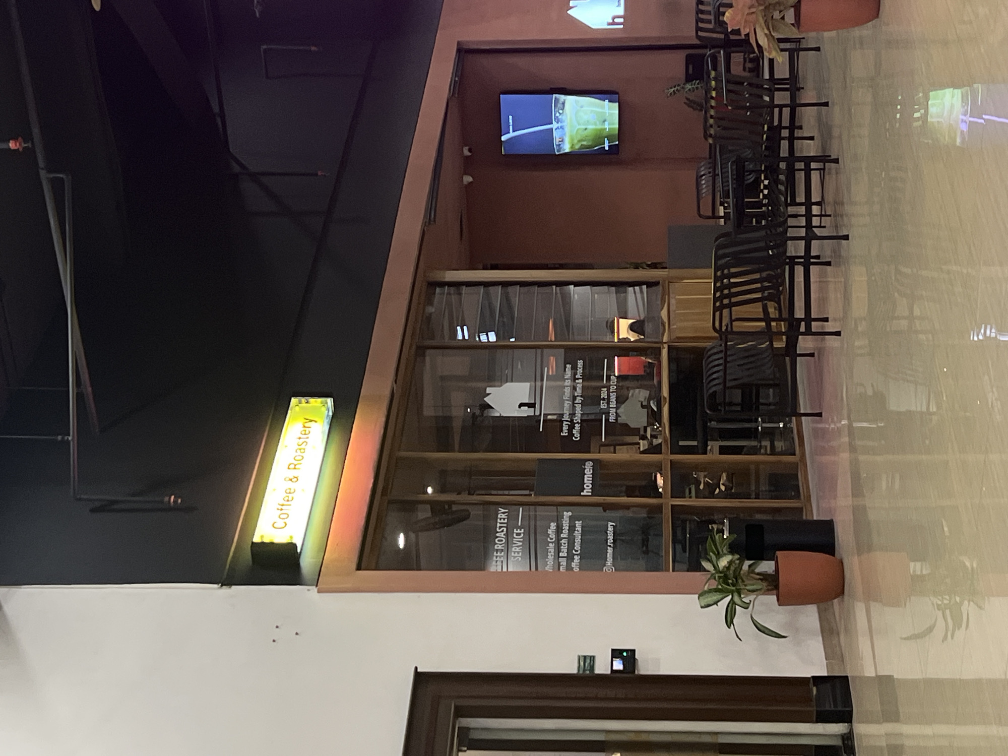 The image shows the exterior of a coffee shop and roastery named "homeie" located in Bandung City, Indonesia. The shop's facade features large glass windows and a prominent illuminated sign that reads "Coffee & Roastery". Inside, the shop appears to be set up with tables and chairs for customers, with some areas visible through the glass. A digital screen displaying a beverage is mounted on the wall to the right. The floor is polished and reflects the interior lights and objects. The overall environment suggests a modern and inviting space for coffee enthusiasts.