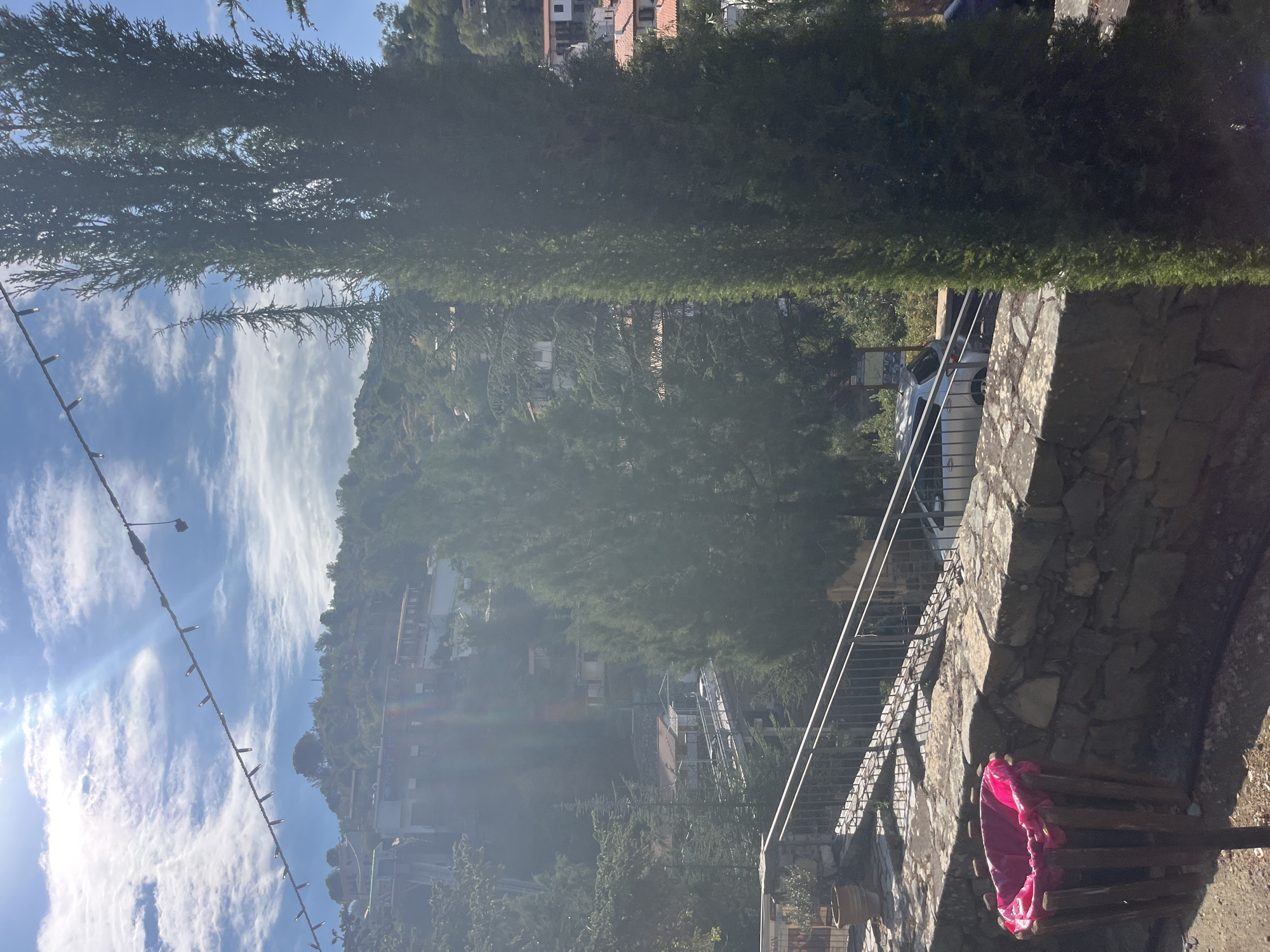 The image depicts a view of Kalopanagiotis, Cyprus, with a focus on the mountainous terrain and village architecture. In the foreground, a stone wall is visible, topped by a wooden trash bin lined with a bright pink plastic bag. To the right of the wall, a tall, slender cypress tree stands prominently. A metal railing lines a pathway or staircase to the left, leading uphill.
In the mid-ground, a road or bridge crosses a valley, with a silver car parked on it. Houses with terracotta roofs are scattered across the green, tree-covered hillsides, typical of the village's setting. The sky is bright blue with scattered white clouds, and the sun casts a strong glare, creating lens flare effects. String lights are suspended across the upper portion of the frame. The overall impression is a sunny, outdoor scene in a hilly, Mediterranean village.