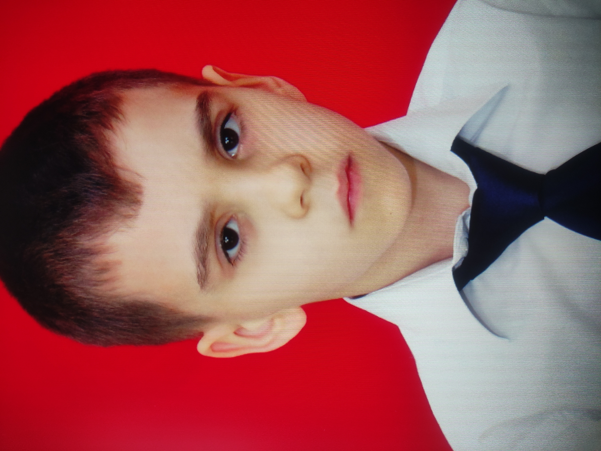 A young boy with dark hair, wearing a white collared shirt and a dark blue tie, is pictured against a solid red background. The boy's head is slightly tilted, and he is looking directly at the camera. His expression is neutral. The image is a headshot, with the boy's shoulders and chest visible. The lighting is even, with no harsh shadows. The red background is evenly lit and appears to be a plain studio backdrop. The image does not provide any information about the location in Baku, Azerbaijan.