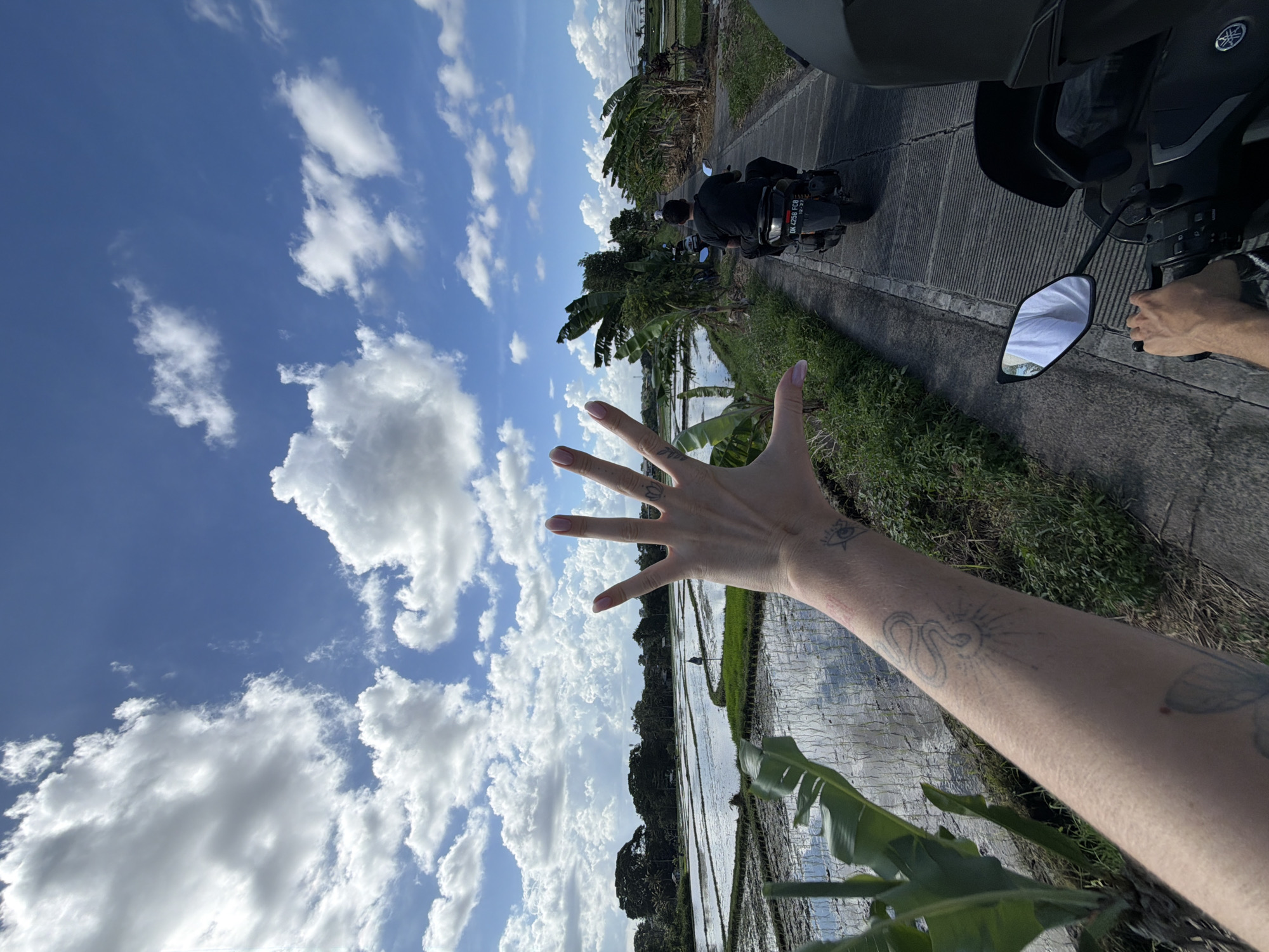 A daytime shot from the back of a moving motorcycle on a road lined with rice paddies in North Kuta, Bali, Indonesia. The sky is blue with scattered white clouds. The left arm and hand of a light-skinned person reach out toward the sky, revealing arm tattoos. Ahead on the road is another person on a motorcycle. The side mirror of the motorcycle the photo is taken from is visible. The road is flanked by green vegetation and rice paddies.