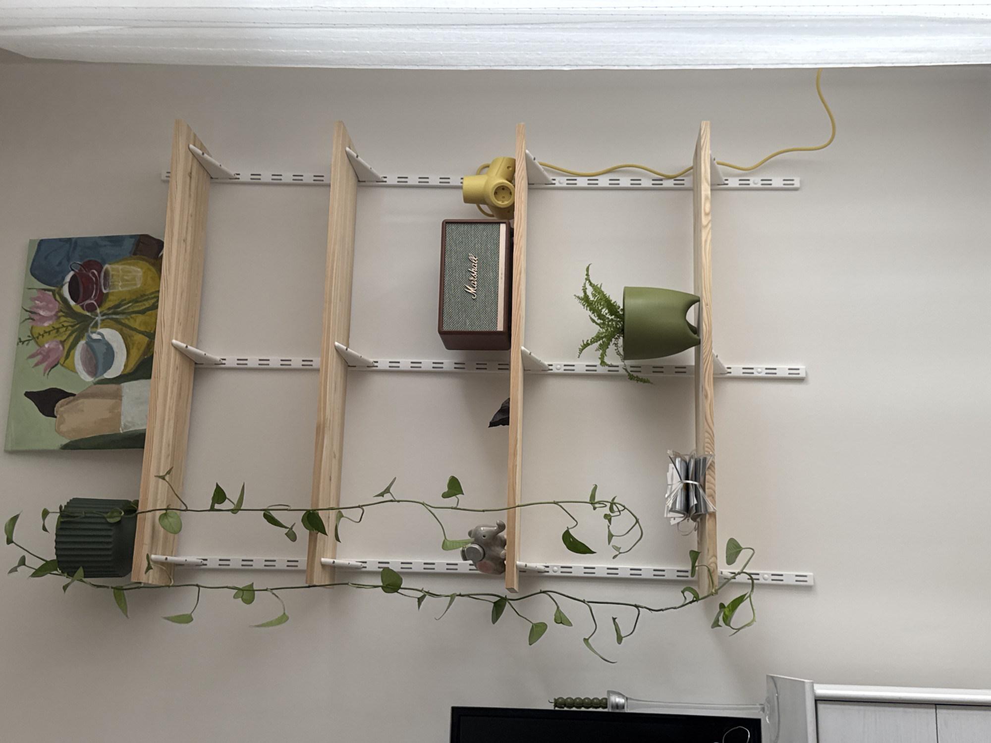 The image shows a wall-mounted shelving unit with four vertical supports made of light-colored wood. The shelves are formed by horizontal metal tracks with slots, attached to the wall. The shelves hold various items: an abstract painting, a dark green ridged planter, a yellow vintage camera, a Marshall speaker, a green watering can with a plant inside, a small gray elephant figurine, and a bundle of objects wrapped in clear plastic. A long, trailing plant is draped across the lowest shelf, with vines extending downwards. Below the shelving unit, the top of a black television and a toy lightsaber are visible. The wall is plain and painted a neutral color. A yellow electrical cord runs along the top of the shelf unit.