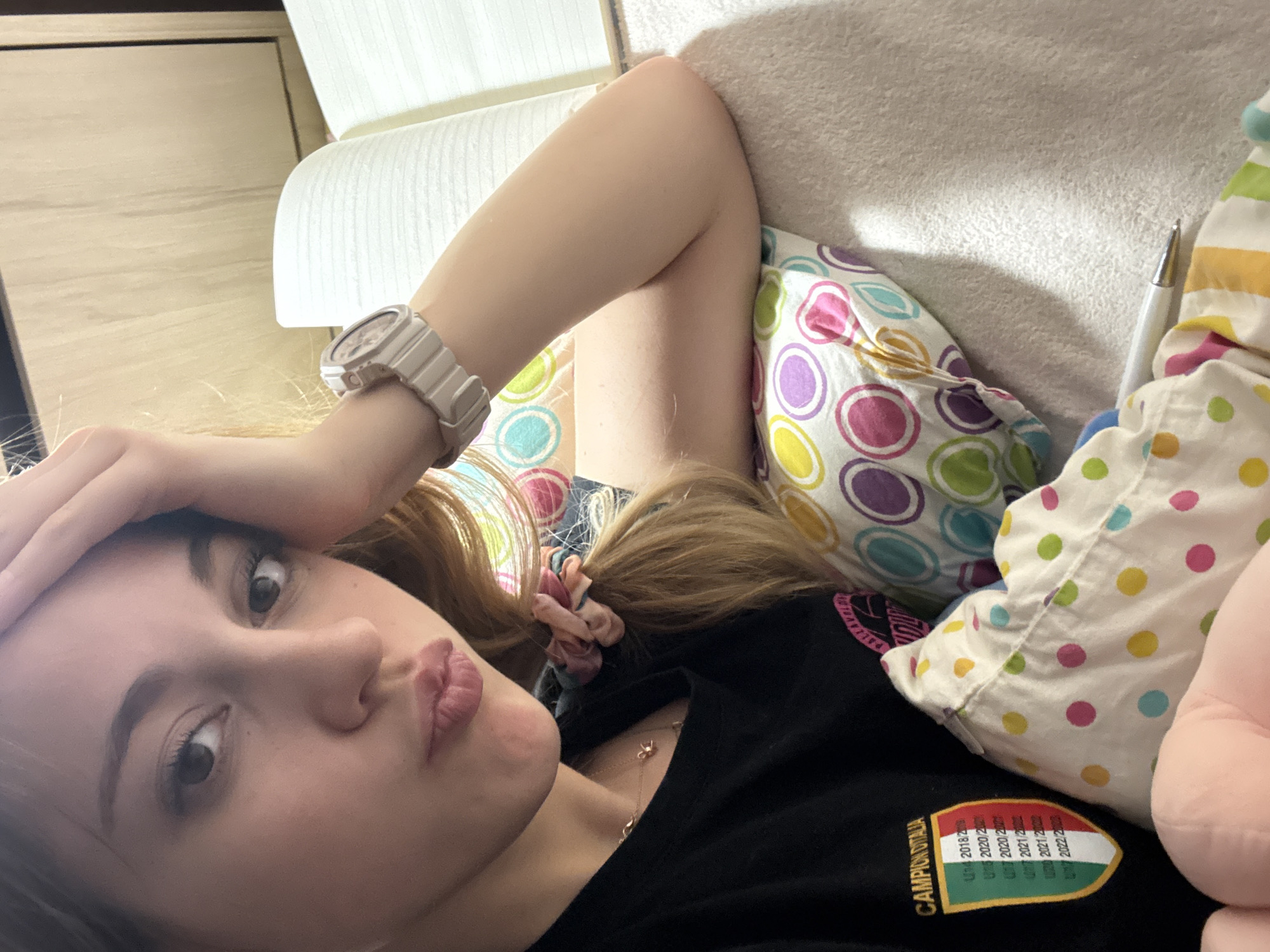 A young woman is lying on a white sofa with a white pillow and two decorative pillows with colorful polka dots. She is wearing a black shirt with a logo that includes the years 2018-2022 and the words "CAMPION D'ITALIA." She has long, light brown hair pulled back with a pink scrunchie. She is wearing a light-colored watch on her left wrist and a gold necklace. Her left hand is resting on her forehead, and she is looking directly at the camera with a pout. The background suggests an indoor environment. The location is not discernible as Sofia, Bulgaria from the image.