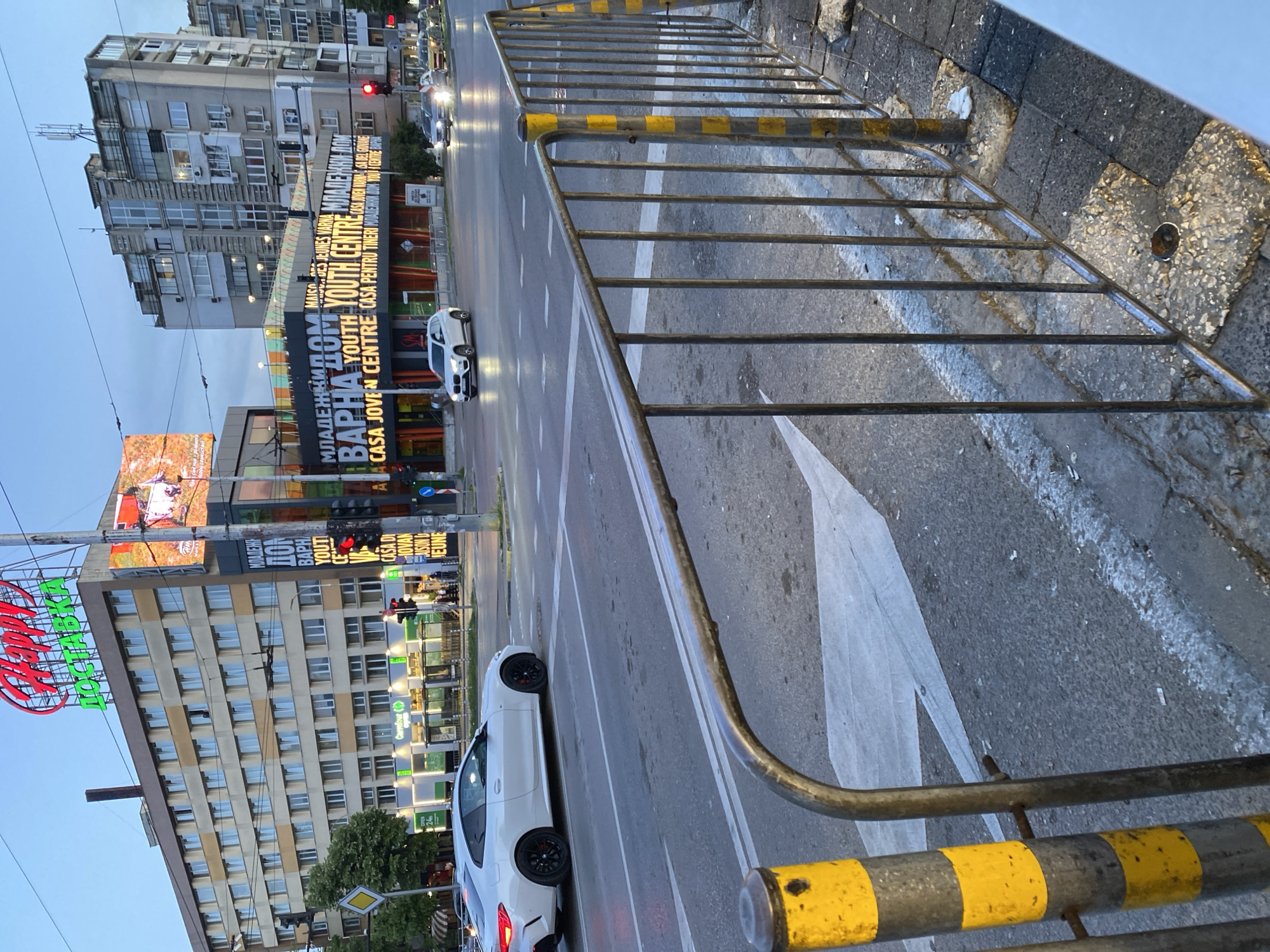 The image presents a street scene in Varna, Bulgaria, viewed from behind a metal safety barrier with yellow and grey stripes. In the background are multi-story buildings, illuminated billboards, and street lighting. The buildings, characteristic of urban residential structures, show a variety of windows. The traffic lights in the distance are visible. The roadway features white lane markings and a white arrow indicating the direction of traffic. A white car is parked on the street, while another one drives in the background. The angle suggests the image was taken from an elevated position. A billboard in the background reads "Mladetzki Dom Varna Youth Centre Casa Joven".