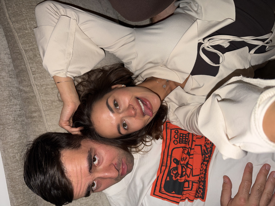 The image depicts an adult male and an adult female positioned closely together, appearing to be lying down on a textured, light beige fabric surface, likely a sofa or bed. The image is taken from an elevated perspective, suggesting a selfie.
The male, positioned on the left, has short dark hair and dark eyes. He wears a white t-shirt featuring a large, stylized orange graphic print of a cartoon character resembling Mickey Mouse within a framed television or radio scene. He looks directly at the camera with a neutral to slightly downturned expression. A small reddish mark is visible below his right nostril. His left hand is partially visible, resting on the lower body area of the female.
The female, positioned on the right, has medium-length wavy dark brown hair and light brown eyes. She smiles broadly at the camera with her tongue extended playfully. She wears a light beige or cream-colored long-sleeved top with wide cuffs, unbuttoned to reveal a dark, possibly black or dark brown, inner garment with lacing details at the front. A delicate silver chain bracelet adorns her left wrist, and a silver-colored necklace with small, geometric pendants is visible around her neck. Her right hand is placed on the male's head, appearing to run through his hair.
The environment is an indoor setting. There are no visual cues within the image to determine a specific location within the city of Tel-Aviv, Israel, or any other geographical area.