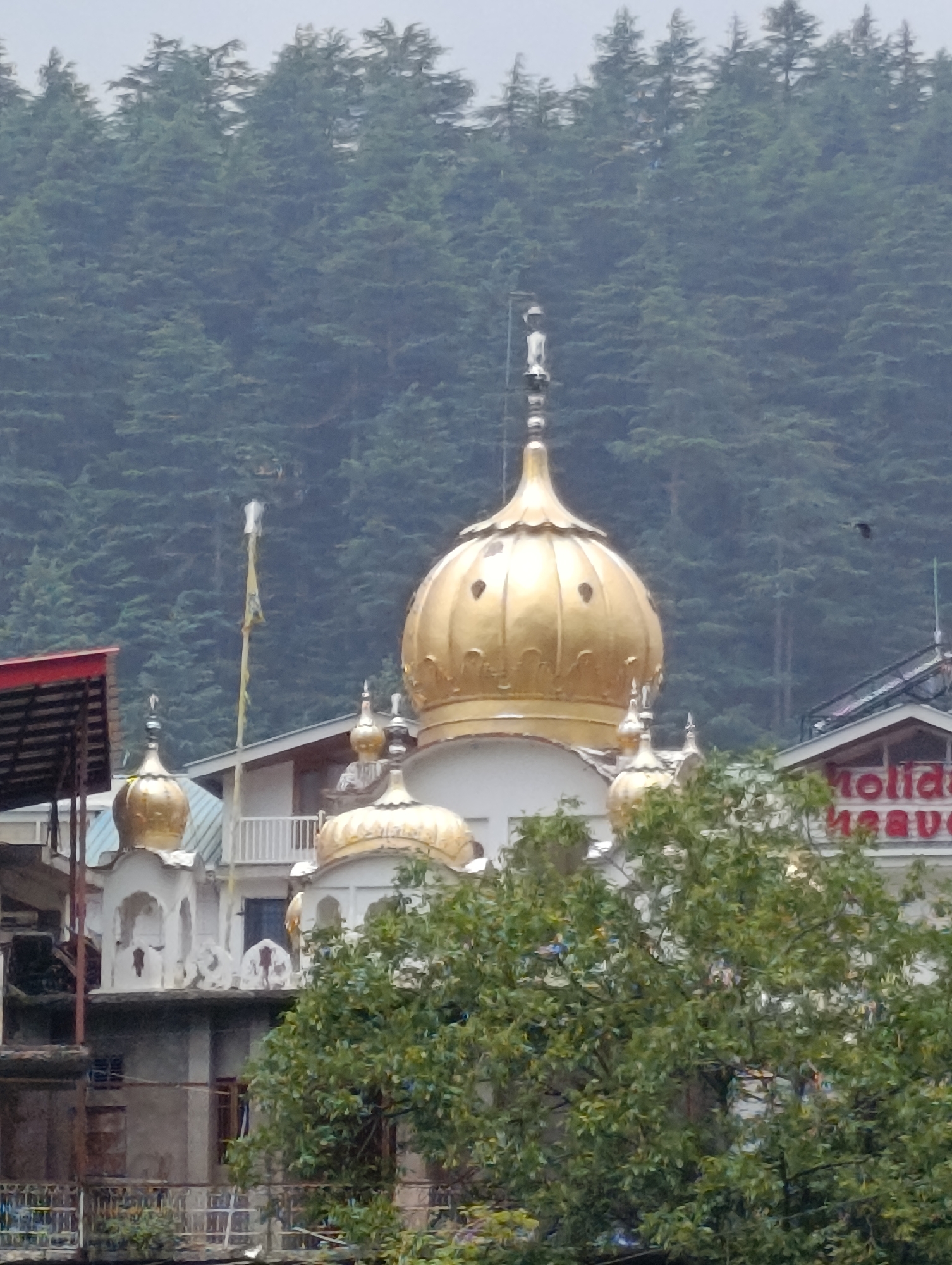 The image depicts a golden dome of a gurdwara against a backdrop of dense green pine trees. The gurdwara features white architecture with ornate detailing and smaller golden domes. A flagpole with a tattered yellow flag stands near the main dome. In the background, a building with the partial text "Holiday Heaven" is visible. The overall scene suggests a mountainous, possibly overcast, environment, typical of Manali, India, a popular hill station in the Himalayas. There are no people or discernible actions captured in the image.