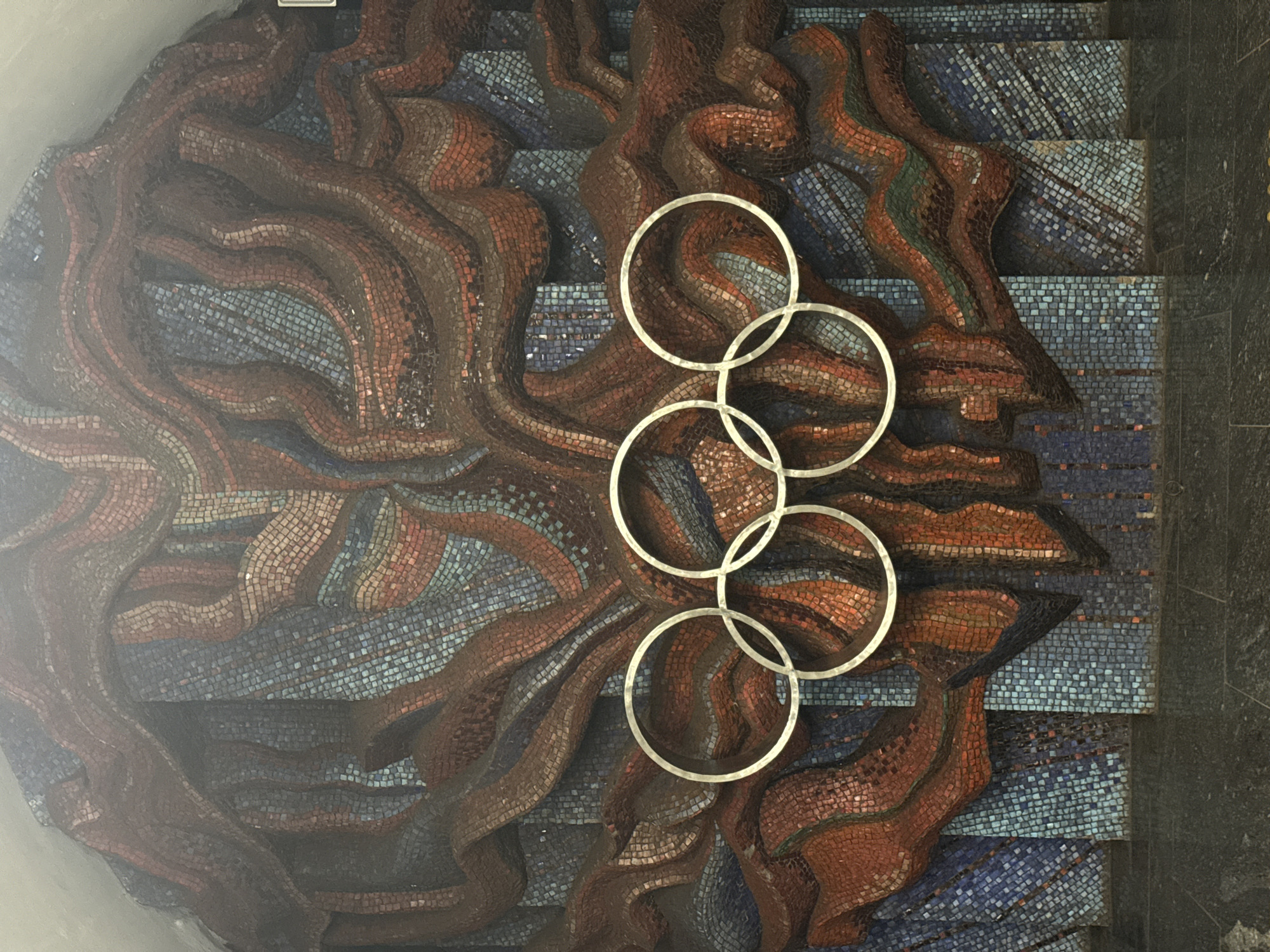 The image shows a mosaic mural depicting abstract forms in red and blue tiles, superimposed with the Olympic rings in a silver metallic finish. The mural is located in the vestibule of the Olimpiiska metro station in Kyiv, Ukraine. The Olympic rings are prominently displayed in the center of the mural, with the bottom rings overlapping. The mosaic art is textured and appears to be made of small, irregularly shaped tiles. The floor in front of the mural is dark and reflective, possibly polished stone or granite. The lighting in the scene is dim, creating a somewhat dramatic effect on the artwork.