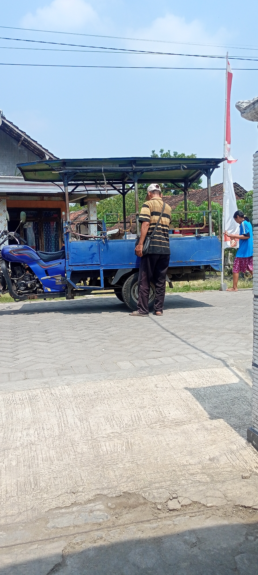 A horizontal, eye-level shot captures a street scene in Gempolklutuk, Indonesia, under a bright, clear sky with a few wispy clouds. The foreground is dominated by a light gray paved surface, with distinct textured lines and some areas of damage and debris.
In the mid-ground, a blue three-wheeled vehicle, likely a modified motorcycle with a cargo bed, is parked on the paved surface. The vehicle has a black canopy stretched over a metal frame above the cargo area. A blue motorcycle is attached to the front of the vehicle.
To the right of the vehicle, a man wearing a horizontally striped polo shirt in yellow and black, dark pants, and a white baseball cap stands facing away from the camera, with a dark cross-body bag slung over his shoulder. Further to the right, near a white pole with a red and white flag partially visible, a person in a blue shirt and patterned shorts is tending to something on a white surface. The background reveals residential buildings with tiled roofs and some greenery. The scene appears to be a street in a village or small town setting.
