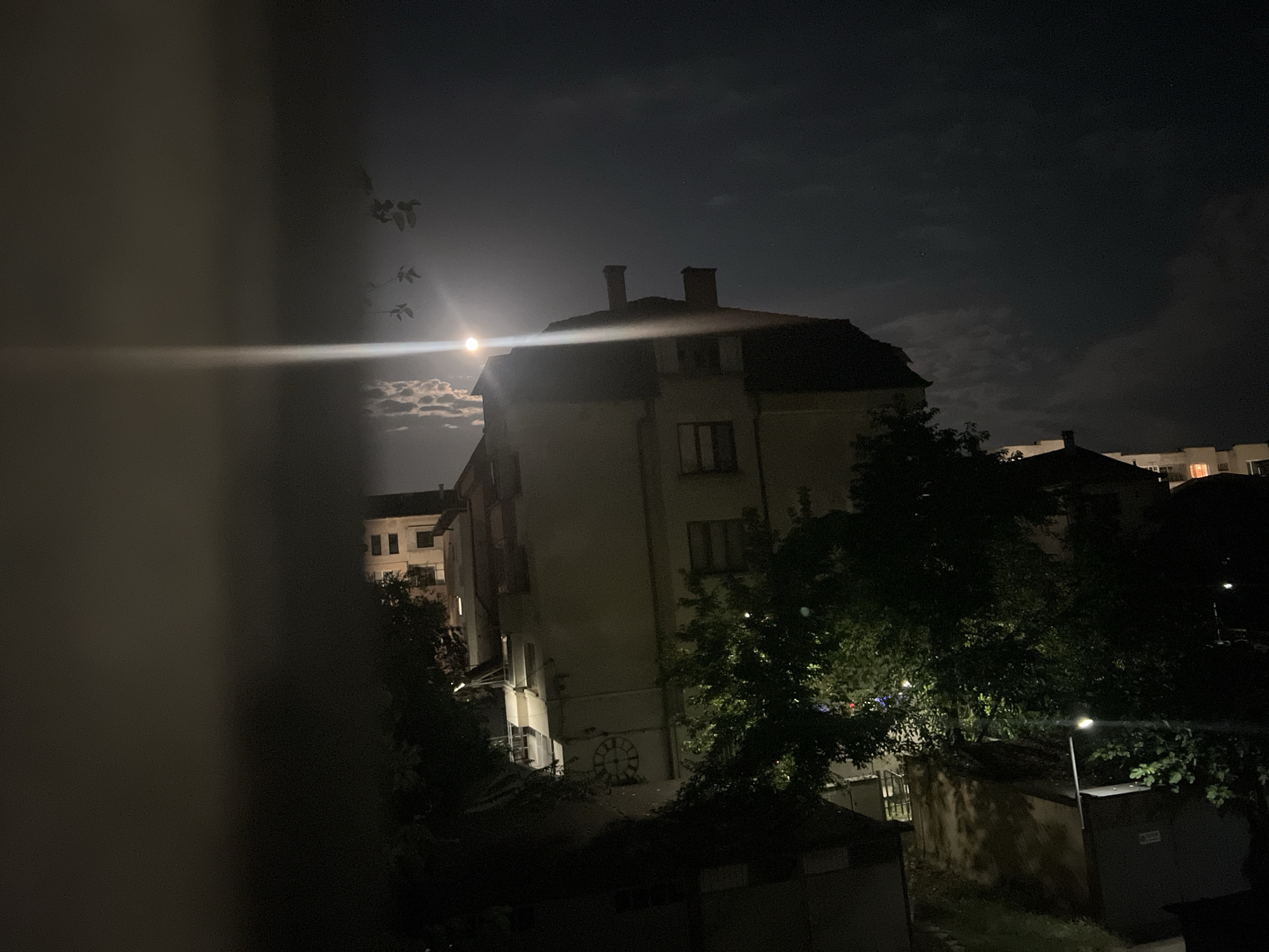 The image captures a nighttime scene in Blagoevgrad, Bulgaria. A large, multi-story building with a dark, sloping roof dominates the center of the frame, illuminated by the moon and possibly artificial light. The building appears to be made of a light-colored material, with visible windows and two chimneys on the roof. To the left of the building, a bright, lens-flared moon is partially obscured by clouds, casting a glow on the scene. A cluster of birds, silhouetted against the sky, are flying towards the left of the frame. In front of the building and to its right, dense foliage from trees obscures parts of the lower levels and surrounding structures. The environment suggests an urban setting, with other buildings visible in the background, some with illuminated windows. A streetlamp is visible to the right of the main building, and a large, circular clock face is mounted on the wall of the central building. The foreground is partially obscured by a dark, out-of-focus element, possibly a window frame or curtain. The overall atmosphere is dark and atmospheric, with a clear indication of nighttime conditions.