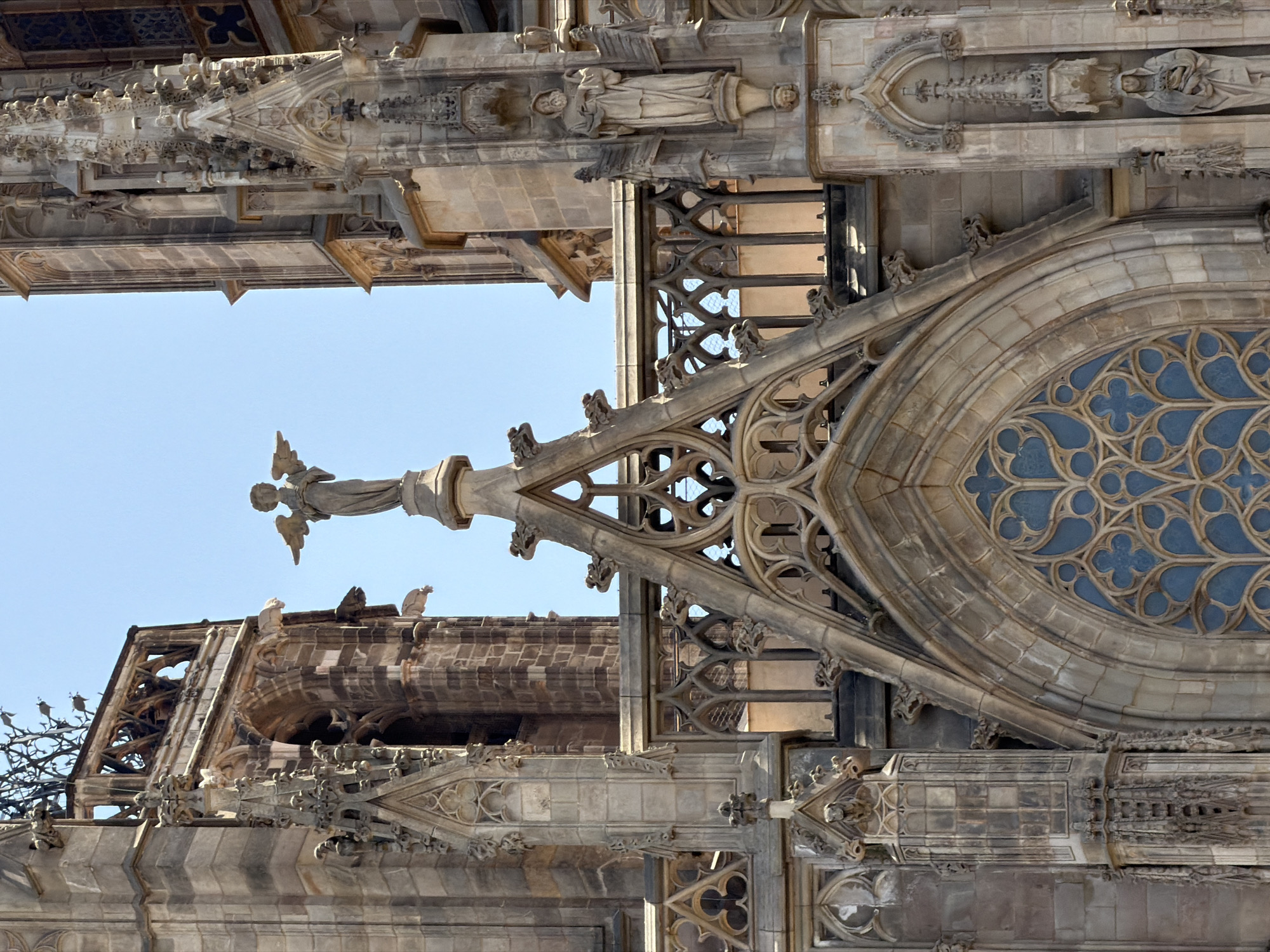 The image shows a detailed view of the upper facade of the Barcelona Cathedral in Barcelona, Spain. The architecture is Gothic, with intricate stone carvings, spires, and arched windows. A statue of an angel with outstretched wings is prominently positioned on a pedestal in the center of the frame. To the right of the angel, another statue depicts a robed figure. Gargoyles are visible adorning the structure. The sky is a clear blue, indicating a sunny day.