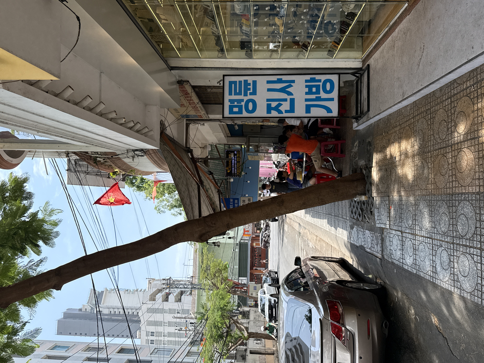 Captured in the Nha Trang Ward of Vietnam, the image presents a street-level view. A tree limb extends horizontally across the frame, partially obscuring the street scene. A Vietnamese flag is visible against the backdrop of urban buildings and power lines. A car is parked along the right side of the street. On the right, people are seated on red plastic stools, near a shop. The shop features a Korean sign. The shop's sign reads “명륜진사갈비” in Korean. The street has a tile pattern, and various signs are visible along the road. The image is taken during daylight.