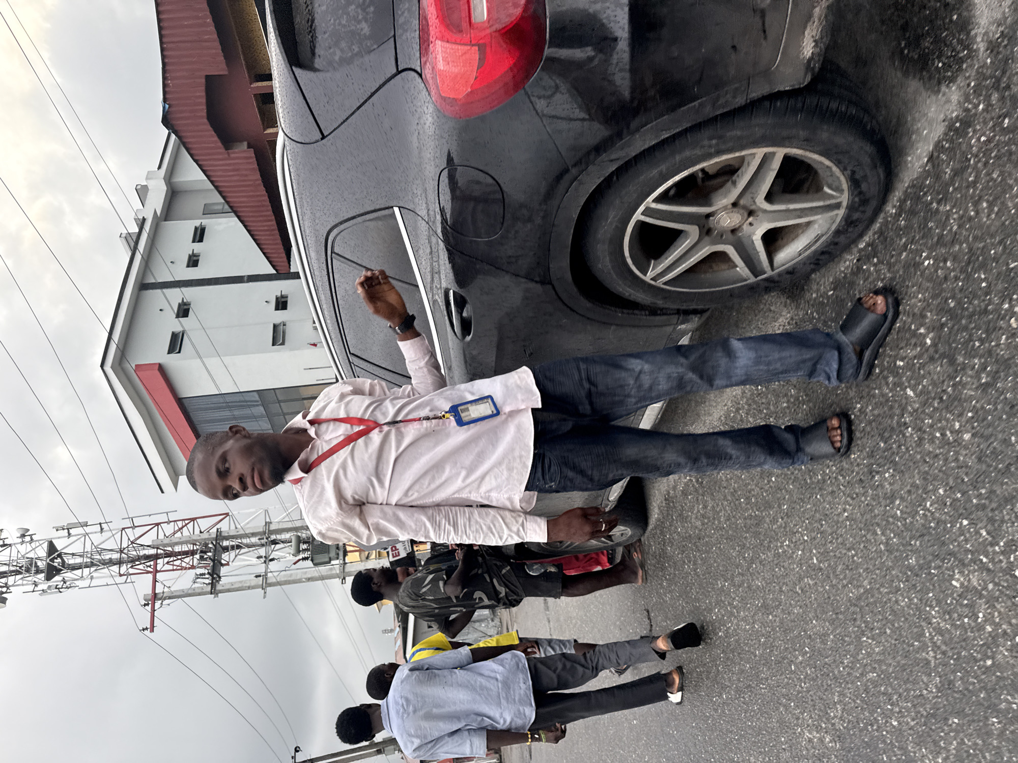 A group of young Black men stand on a wet asphalt road next to a black SUV. The man in the foreground, wearing a light pink button-down shirt, dark jeans, and black sandals, leans against the car. He has a red lanyard with a blue ID badge around his neck. Behind him, another man in a light blue shirt and dark pants walks away from the camera, while a third man in a camouflage t-shirt and dark shorts stands facing the SUV. Further back, a man in a yellow shirt is visible. The background shows a modern, multi-story building with red accents and a large cell tower. The sky is overcast. The scene appears to be in Iranla, Nigeria.