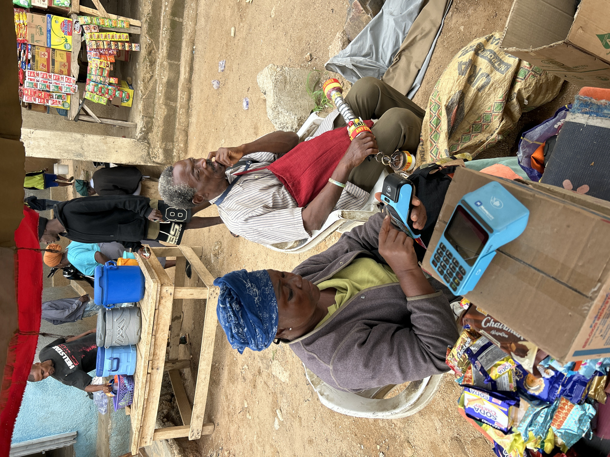 In an outdoor setting in Jos, Nigeria, an elderly woman wearing a blue headscarf and a grey jacket is seated and appears to be operating a blue electronic payment terminal. The terminal rests on a cardboard box laden with assorted packaged snacks and candies.
Nearby, an elderly man, also seated, holds a decorated, possibly ceremonial, pipe-like object. He wears a striped shirt and a red satchel. To the left of the frame, other people are partially visible, including a man in a "HELLSTAR" t-shirt. A wooden structure, likely a makeshift shop, displays an array of packaged goods and drinks. The environment appears to be a bustling marketplace or street vending area.