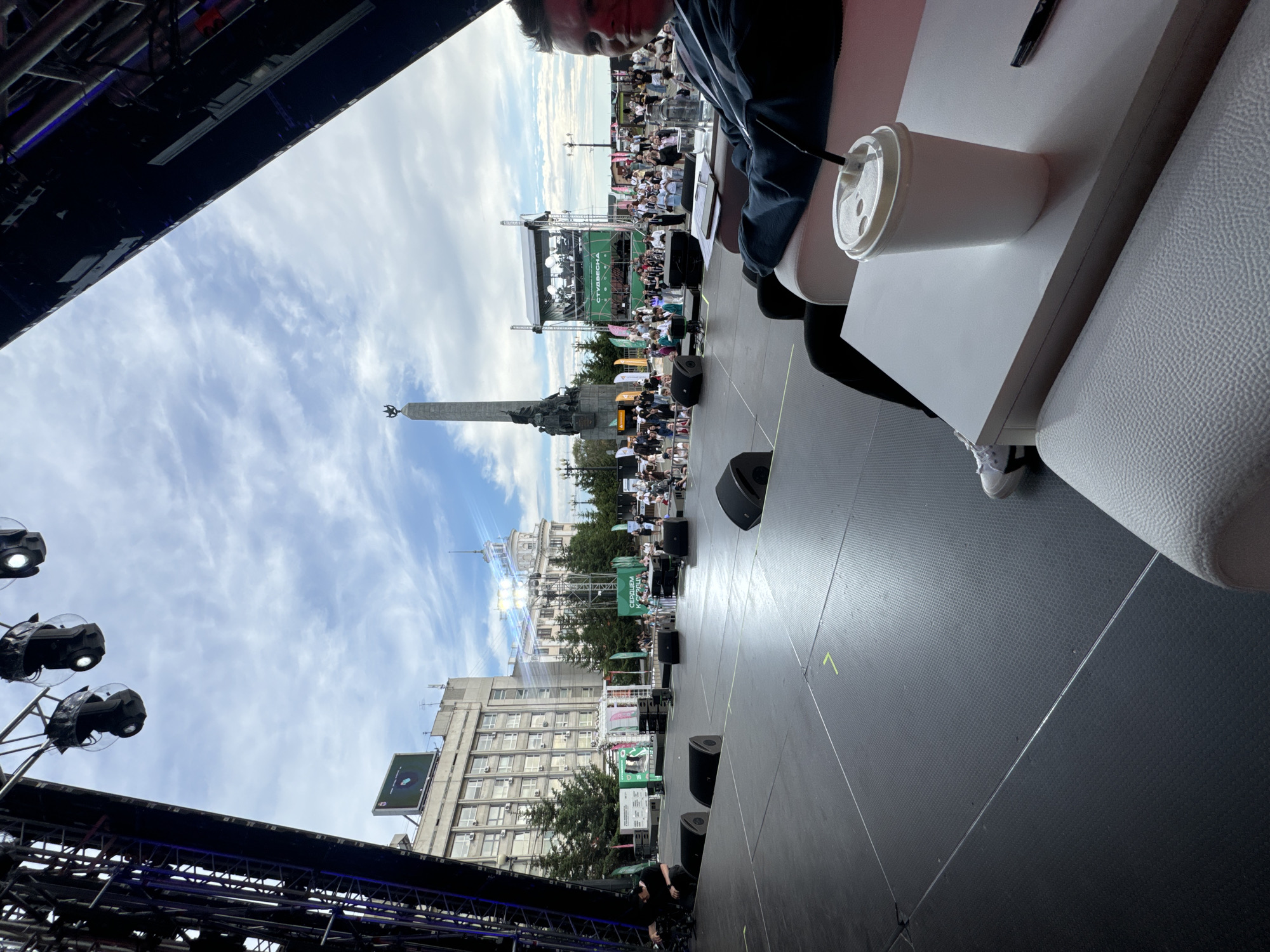 The image shows a wide view of an outdoor stage setup in the city of Khabarovsk, Russia. A large stage is in the foreground, with black speaker boxes scattered across its surface. In the background, a crowd of people are gathered, and a stage with a large banner that reads "СТУДВЕСНА" is visible. To the left of the crowd, a tall obelisk monument stands, a prominent landmark in the city. The sky is partly cloudy. A person is seated on a white couch in the foreground, with a cup and a pen on a white table in front of them. The overall scene suggests an outdoor event or festival is either about to begin or has just concluded.