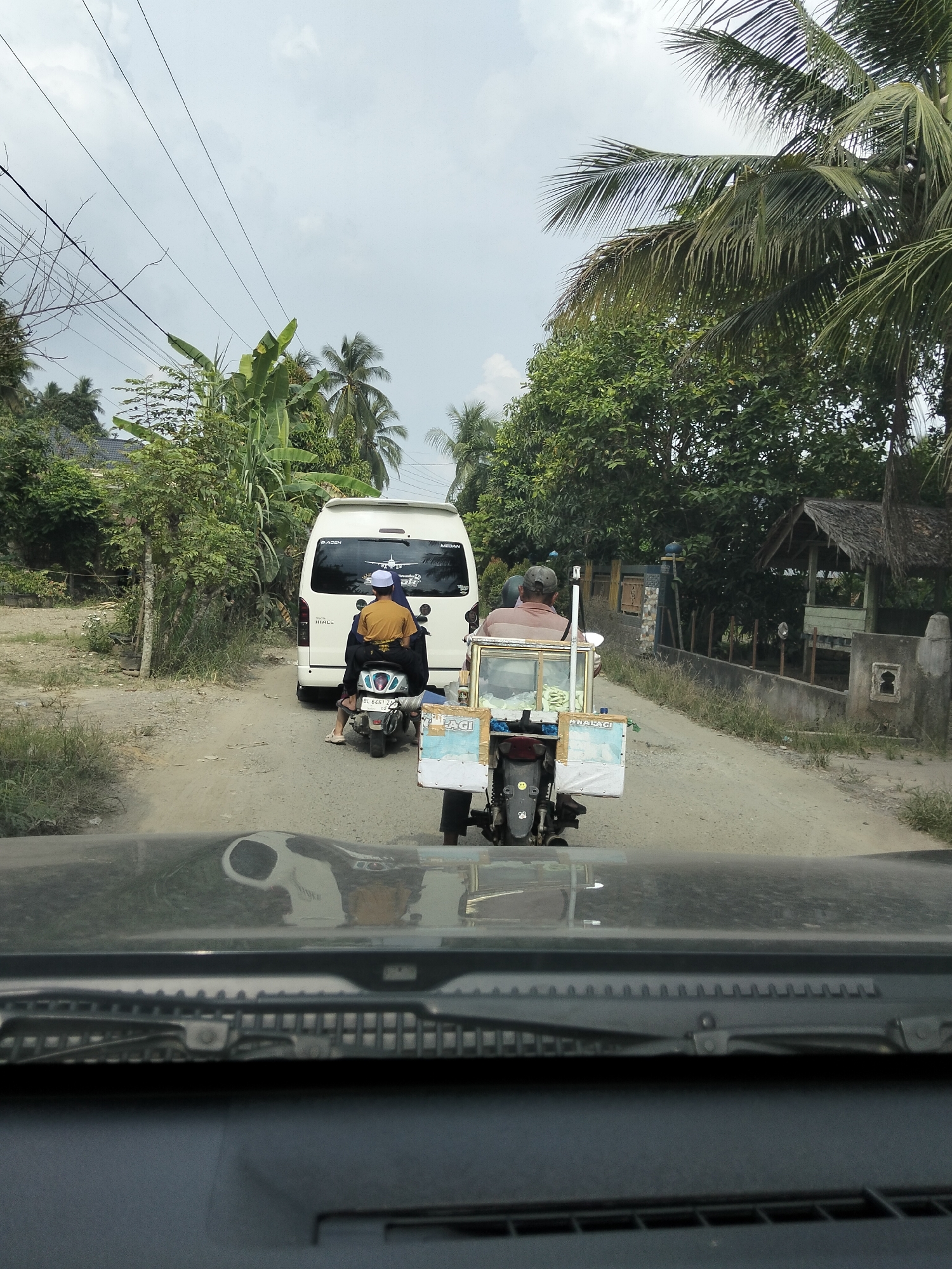 In Teupinreudeup, Indonesia, the image captures a scene on a dirt road with a white van and two motorcycles. The van, a Toyota Hiace, is parked on the road, with two people on a motorcycle behind it. The driver of the motorcycle is wearing a light brown shirt and a grey cap, while the passenger wears a yellow shirt and a white cap. Another motorcycle with a sidecar carrying what appears to be food items is positioned in front of the van. The sidecar has white panels with text, and the motorcycle is ridden by a person wearing a light brown shirt and a grey cap. The environment is rural, with lush green trees and vegetation lining the road. The sky is overcast. The photograph is taken from inside a vehicle, showing the dashboard and windshield.