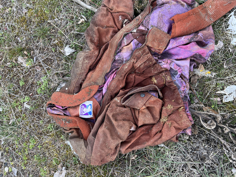 A heavily soiled, crumpled, reddish-brown suede-like jacket with a rust-colored ribbed knit collar and cuffs lies discarded on sparse, dry grassy ground mixed with dirt, twigs, and plant debris in Stoianovo, Bulgaria. The jacket is unzipped, revealing a vibrantly patterned purple garment with pink and orange abstract designs underneath. A blue brand label affixed to the interior collar of the jacket displays the white text "YESSICA" above a white silhouette of a woman's head. The jacket's exterior shows substantial dirt and plant matter accumulation, particularly on the ribbed collar and lower sections. A single flap pocket with a dark, circular snap button is visible on the jacket's lower left side. One rust-colored ribbed cuff is visible detached or torn from the jacket at the bottom right. No individuals are present.