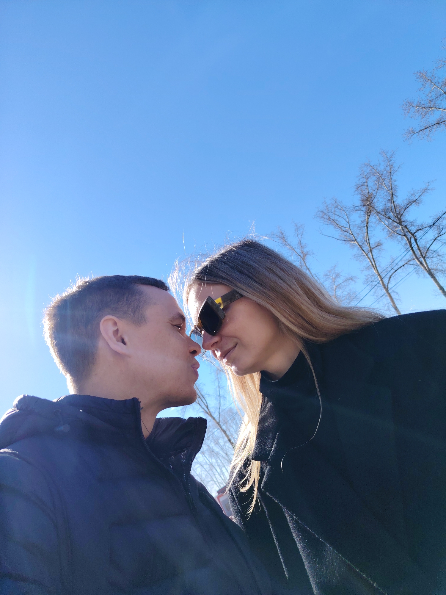 A couple, a man and a woman, are standing close together with their faces near each other, possibly about to kiss. The man is wearing a dark, puffy jacket with a hood. The woman has long blonde hair and is wearing sunglasses and a dark coat. The background is a clear blue sky with some bare trees visible in the upper right corner. The location is most likely in Chaikovsky, Russia, given the prompt.