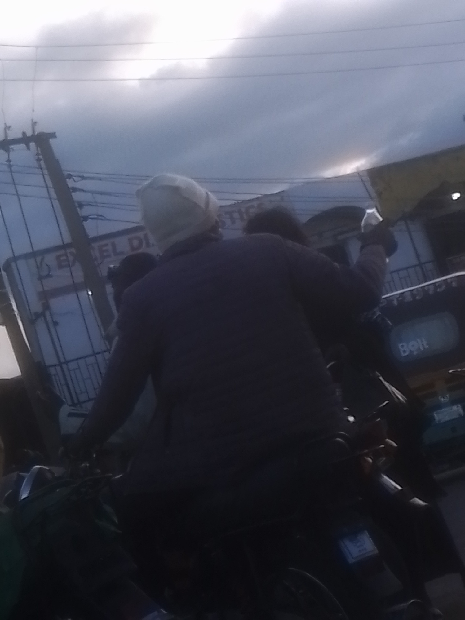 In Gwong, Nigeria, a person wearing a white head covering rides on the back of a motorcycle. The rider is wearing a dark jacket and appears to be holding on to the driver. The background shows a cloudy sky and a street scene with buildings and electrical poles. One of the buildings has a sign that reads "EXCEL DI STIC". Another vehicle, possibly a truck, is visible with the word "Bolt" in a white rectangle. The scene is dimly lit, suggesting it might be dusk or overcast.