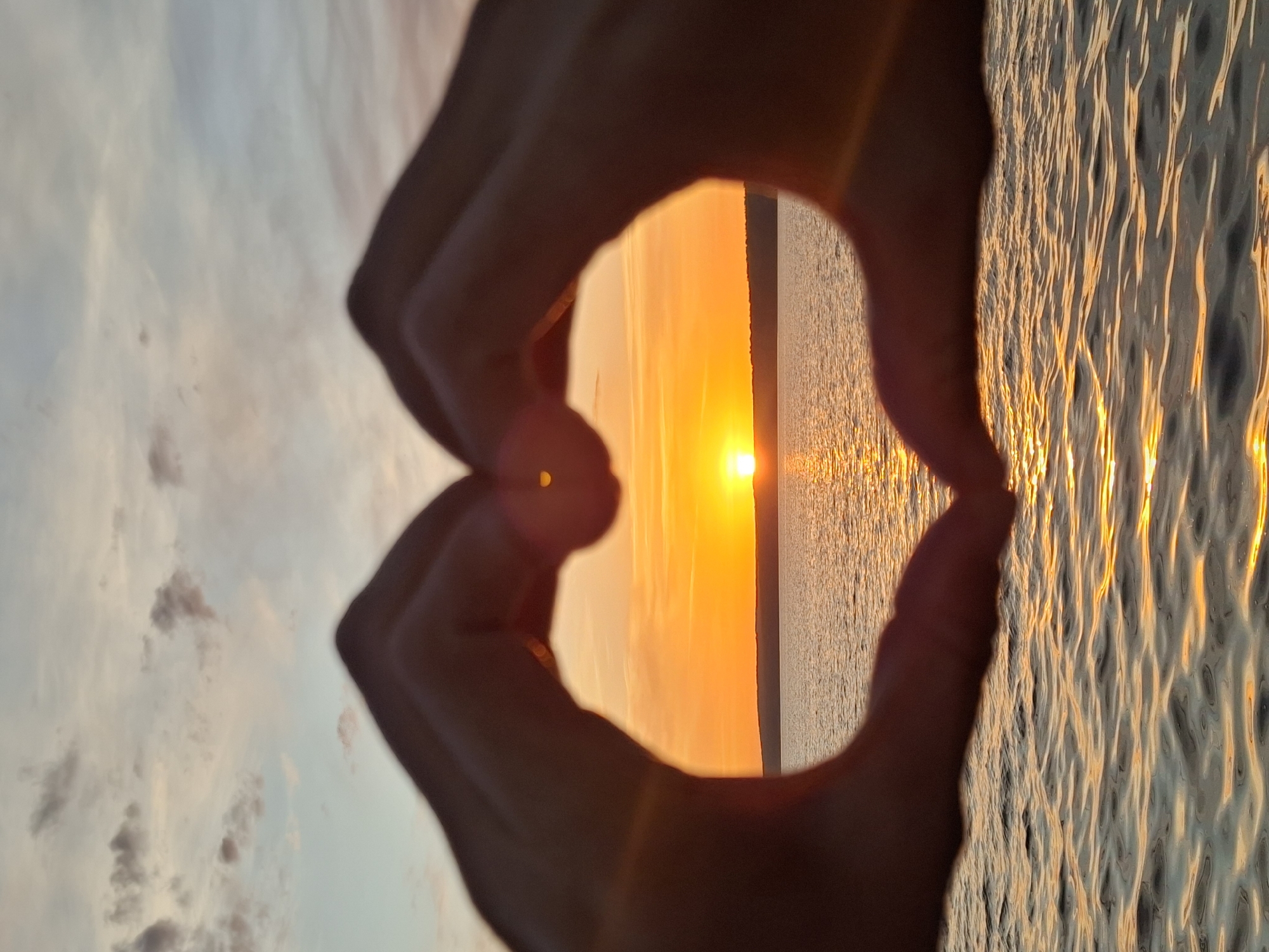 Captured in Chaikovsky, Russia, the image features a sunset over water, framed by two hands forming a heart shape. The sun is positioned at the horizon, its light reflecting on the water's surface creating a shimmering effect. The sky above displays hues of orange, yellow, and faint blues, suggesting either dawn or dusk. The hands are silhouetted against the bright sun and sky, obscuring their features. The water appears calm, with subtle ripples reflecting the sunset's golden light.