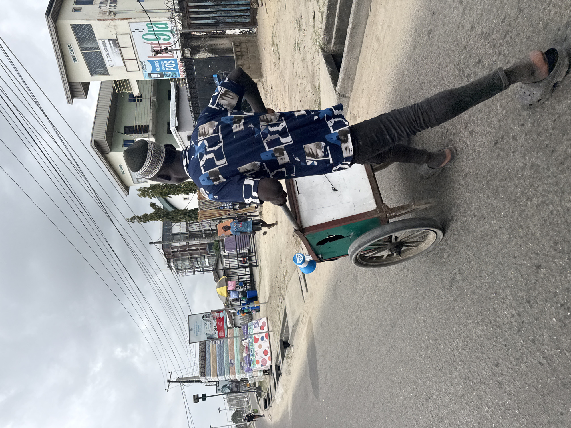 The image depicts a man pushing a cart along a street in Ado, Nigeria. The man is wearing a blue patterned shirt, dark trousers, and a patterned cap. He is pushing a two-wheeled cart that has a green body and a white front panel. The cart has a blue object attached to its front. The street appears to be paved, with a gutter running alongside it. In the background, there are buildings, some of which appear to be shops or offices. One building is labeled "TIMLOT COMPLEX". There are also power lines visible overhead. Other people and merchandise are visible in the background, suggesting a commercial area or market. The sky is overcast, indicating a cloudy day.