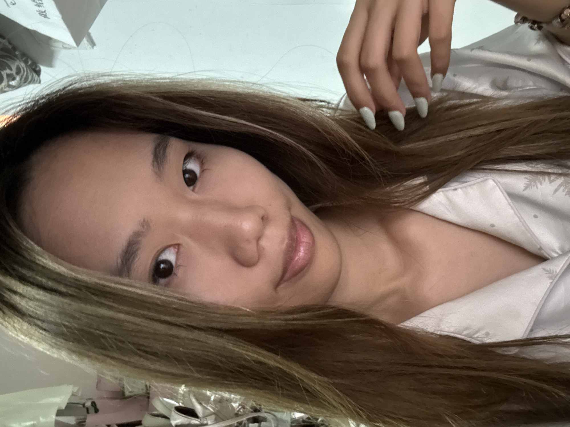 A young woman with brown hair and light green nail polish is lying down, looking at the camera. She is wearing a light-colored pajama top with a subtle pattern. The background is blurred, but includes what appears to be a desk or cluttered surface with papers and other items. The location within the city of Nan'an District, China is not discernible from the image.