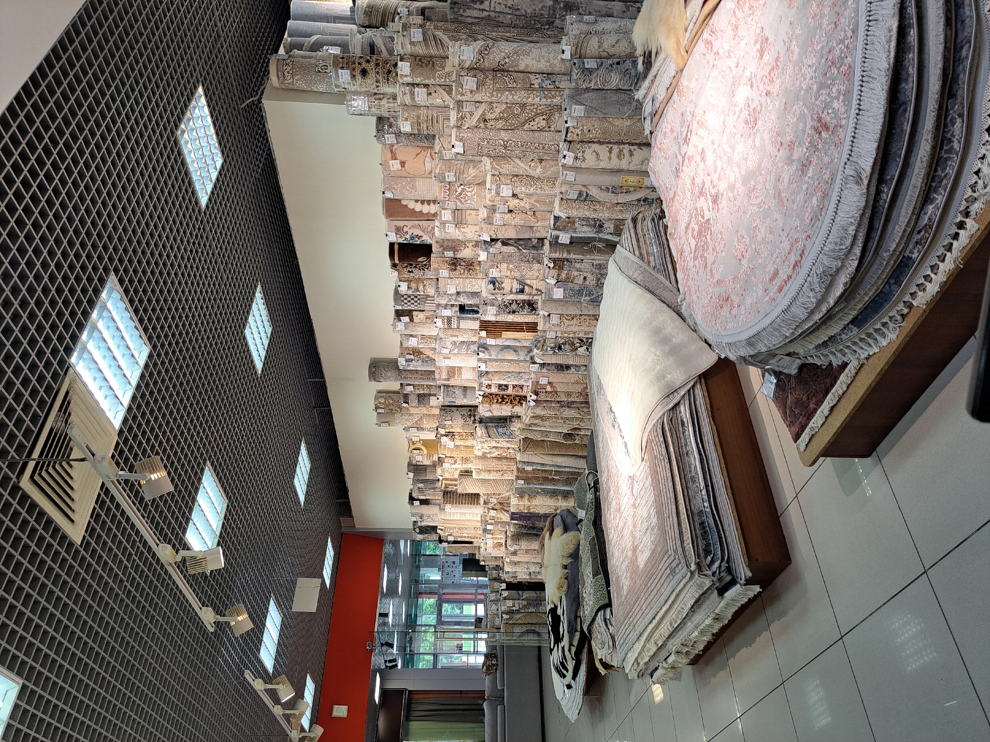 The image shows the interior of a rug store. The store is well-lit with fluorescent lights in a grid ceiling. The walls are white, and the floor is tiled in a light color. The store is filled with a large selection of rolled and folded rugs of various sizes, patterns, and colors. In the foreground, there are stacks of folded rugs, and to the right, a round display of folded rugs with fringe. In the background, there are tall displays of rolled rugs. Some of the rugs are wrapped in plastic. In the far background, through a glass partition, there is a glimpse of the outdoors with trees and a building with large windows, suggesting it might be a shopping center. There are no people visible in the image. The store is located in the city of Barnaul, Russia.