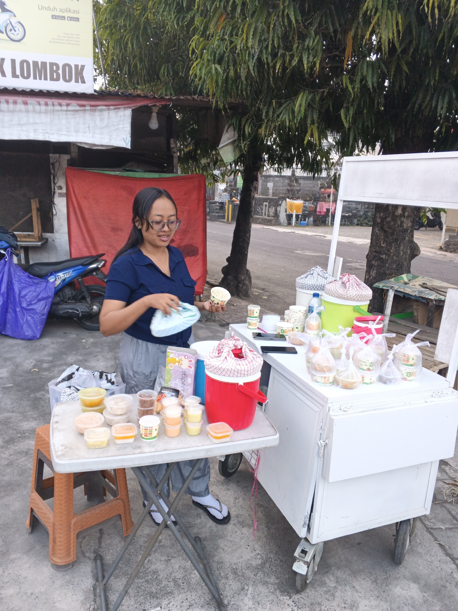 A woman stands behind a food cart in Sukawati, Indonesia, arranging food items for sale. The cart, a white wooden structure with wheels, is positioned on a paved area beside a street. On the cart's countertop, numerous small containers of food are displayed, many of which are individually wrapped in plastic bags. A large red cooler sits on the left side of the cart, and a green cooler is visible on the right. The woman, wearing a dark blue short-sleeved shirt and grey pants, is holding a small white container and a green and white patterned cloth. A small plastic stool is placed to the left of the cart, holding more food containers. In the background, a blue motorcycle is parked, and a red tarp hangs behind it. Trees with lush green foliage are visible above the scene. A street is visible in the far background with a few buildings and trees. The overall lighting suggests it is daytime, possibly late afternoon.