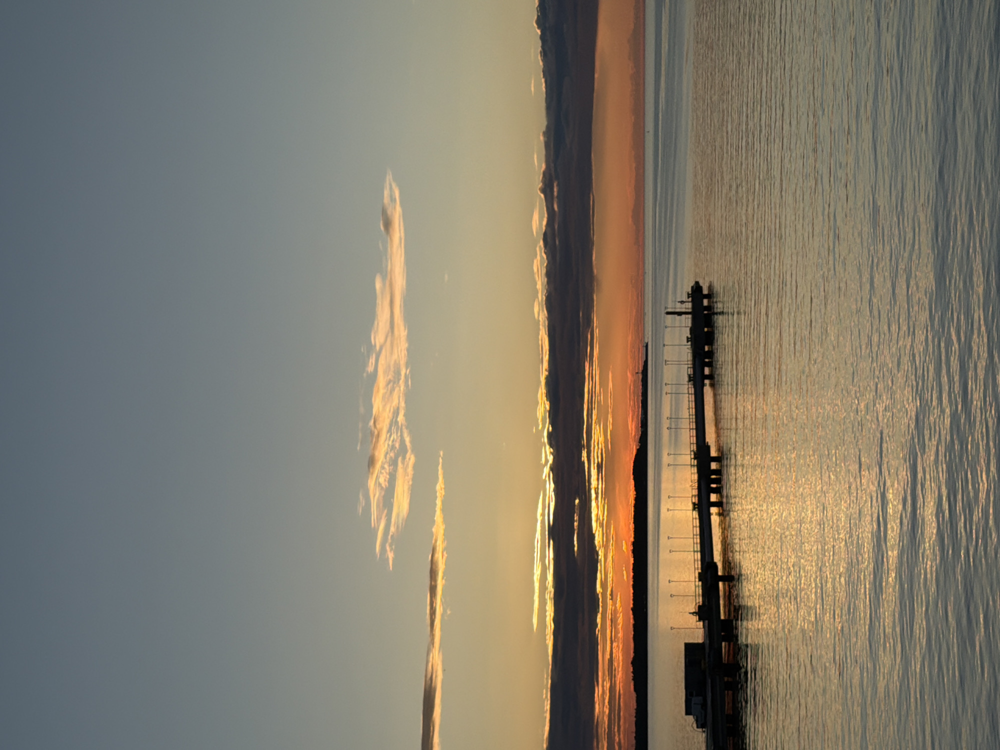 The image shows a view of the sea and a pier at sunset. The sky is clear with a few clouds. The sun is setting, casting an orange glow over the water. There is a pier in the foreground with lights. The location is likely in the city of Tallinn, Estonia, as suggested by the presence of a pier that could be part of the city's coastline. The scene appears to be tranquil and serene.
