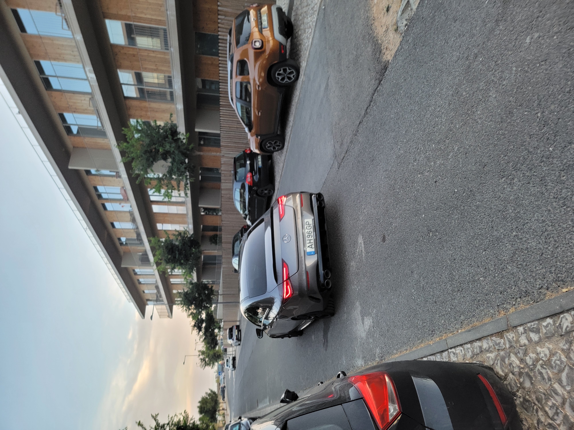 The image depicts a street scene in Quarteira, Portugal, with a modern, multi-story building on the right. The building has wooden paneling and multiple floors with balconies and large windows. Several cars are parked or driving on the asphalt road in front of the building.
In the foreground on the left is the rear of a black car, showing its red taillight and part of its trunk. To the right of this car is a grey Mercedes-Benz sedan with its rear facing the camera. This car has a license plate reading "AH96GP". Further down the road, a dark brown Dacia Duster SUV is parked. Behind the Mercedes, a black Volkswagen Golf hatchback is also parked. Other cars are visible further down the street, suggesting a residential or commercial area.
The street is paved with asphalt, and the right side of the road is lined with cobblestone and a low concrete curb. Trees with green foliage are planted along the sidewalk and in front of the building. The sky above is light blue with some wispy clouds, indicating it is likely daytime, possibly late afternoon or early evening given the lighting. There are streetlights visible along the road. No people are clearly visible in the image.