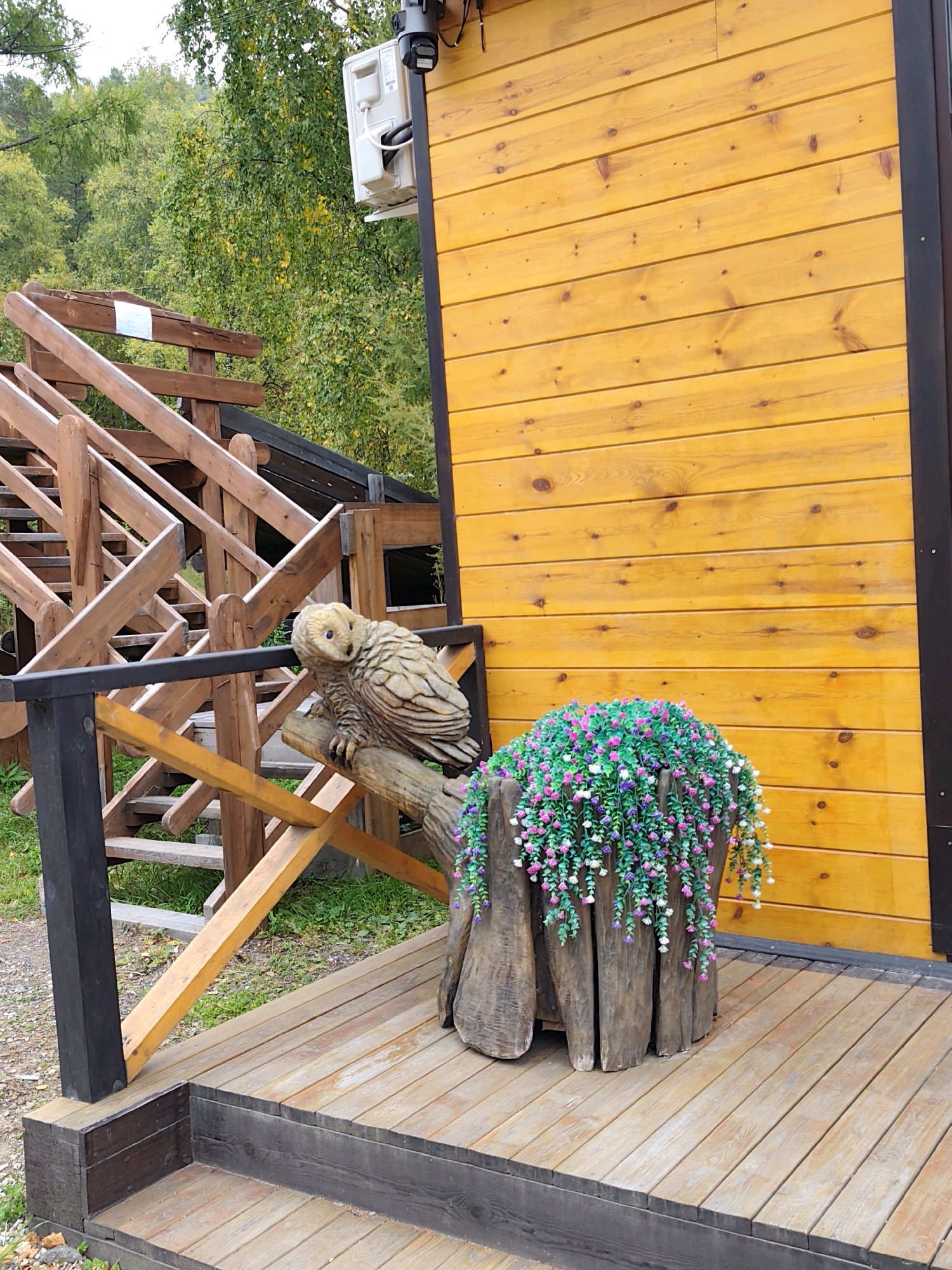 The image captures a wooden deck leading to a bright yellow wooden building. To the left, a wooden staircase with dark gray posts ascends. On the deck, near the building, is a rough-hewn wooden stump adorned with a cascade of artificial pink and white flowers. A carved wooden owl is perched on a railing in front of the stump. In the background, lush green trees and vegetation suggest a natural, possibly rural or park-like setting. A white air conditioning unit and a black security camera are visible mounted on the side of the building. The lighting suggests an overcast day. The scene appears to be outdoors, in a location that combines rustic charm with modern amenities.