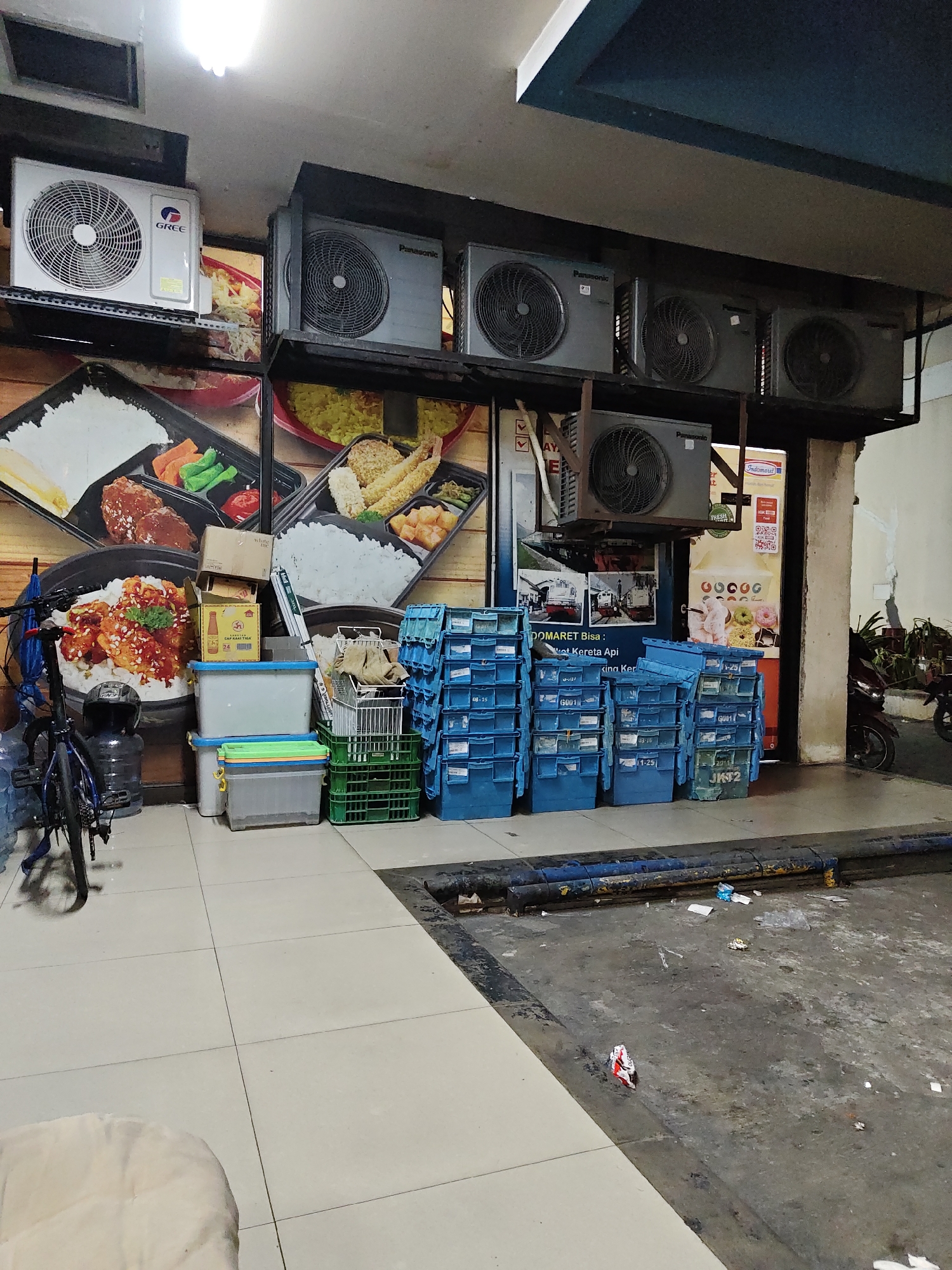 The image depicts an outdoor area, likely behind a commercial establishment, in North Jakarta, Indonesia. The scene is cluttered with various objects. In the background, several air conditioning units are mounted on shelves and on a higher ledge. Below these units, large posters featuring food items and potentially transportation (trains) are displayed on a wall. In the foreground, a collection of blue and green plastic storage bins are stacked high. To the left, a bicycle with an umbrella is leaning against a wall, accompanied by several large water jugs. A few cardboard boxes and other miscellaneous items are also present. The ground is a mix of tiled flooring and a rougher, grey concrete surface with scattered debris. The overall impression is one of a storage or service area that is functional but untidy.