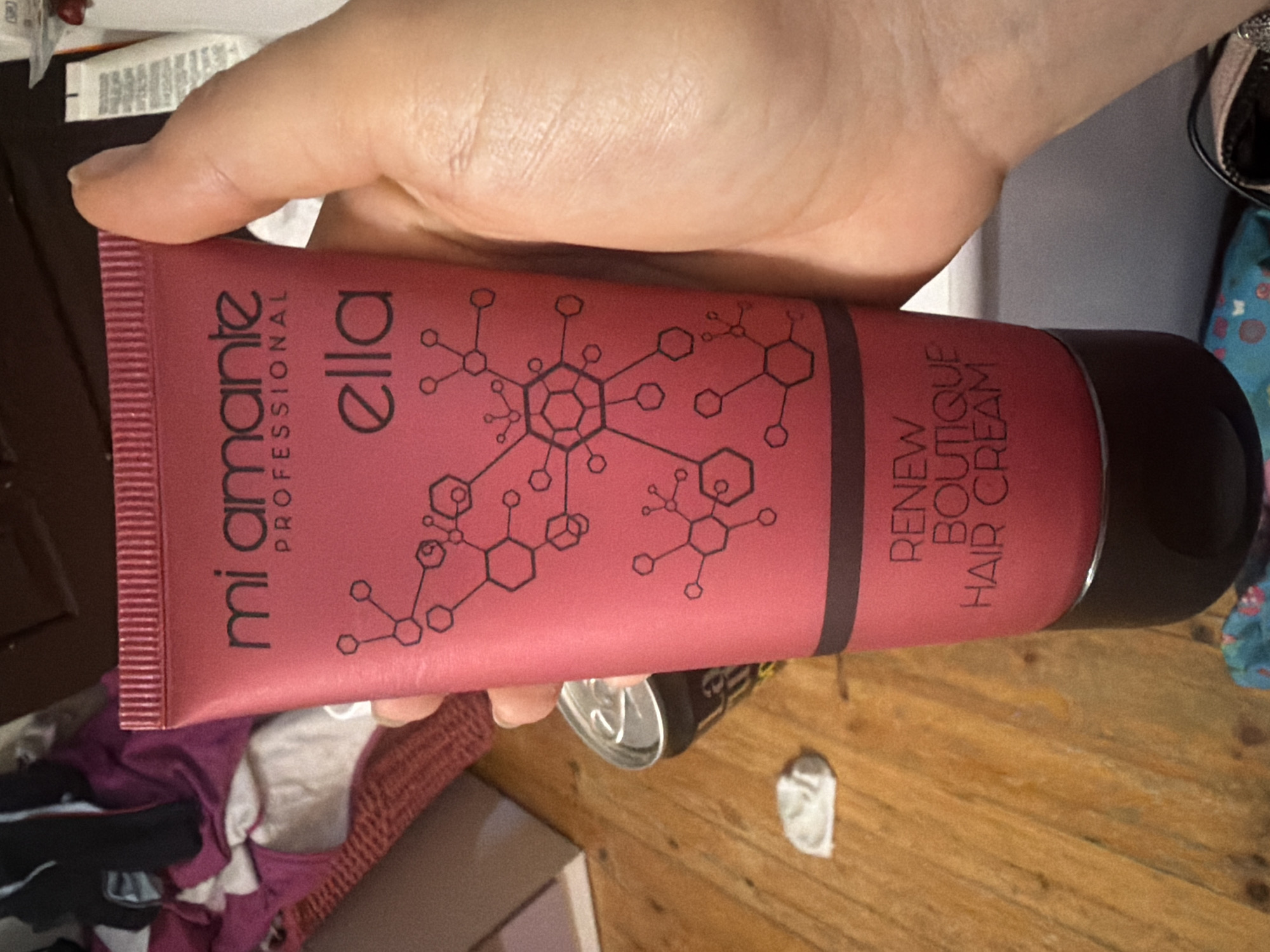 A hand holds a red tube of "mi amante ella professional" Renew Boutique Hair Cream with black molecular-like patterns, over a wooden surface with a soda can and a crumpled tissue. The background includes clothing items and other miscellaneous objects. The location is Unknown, Unknown.