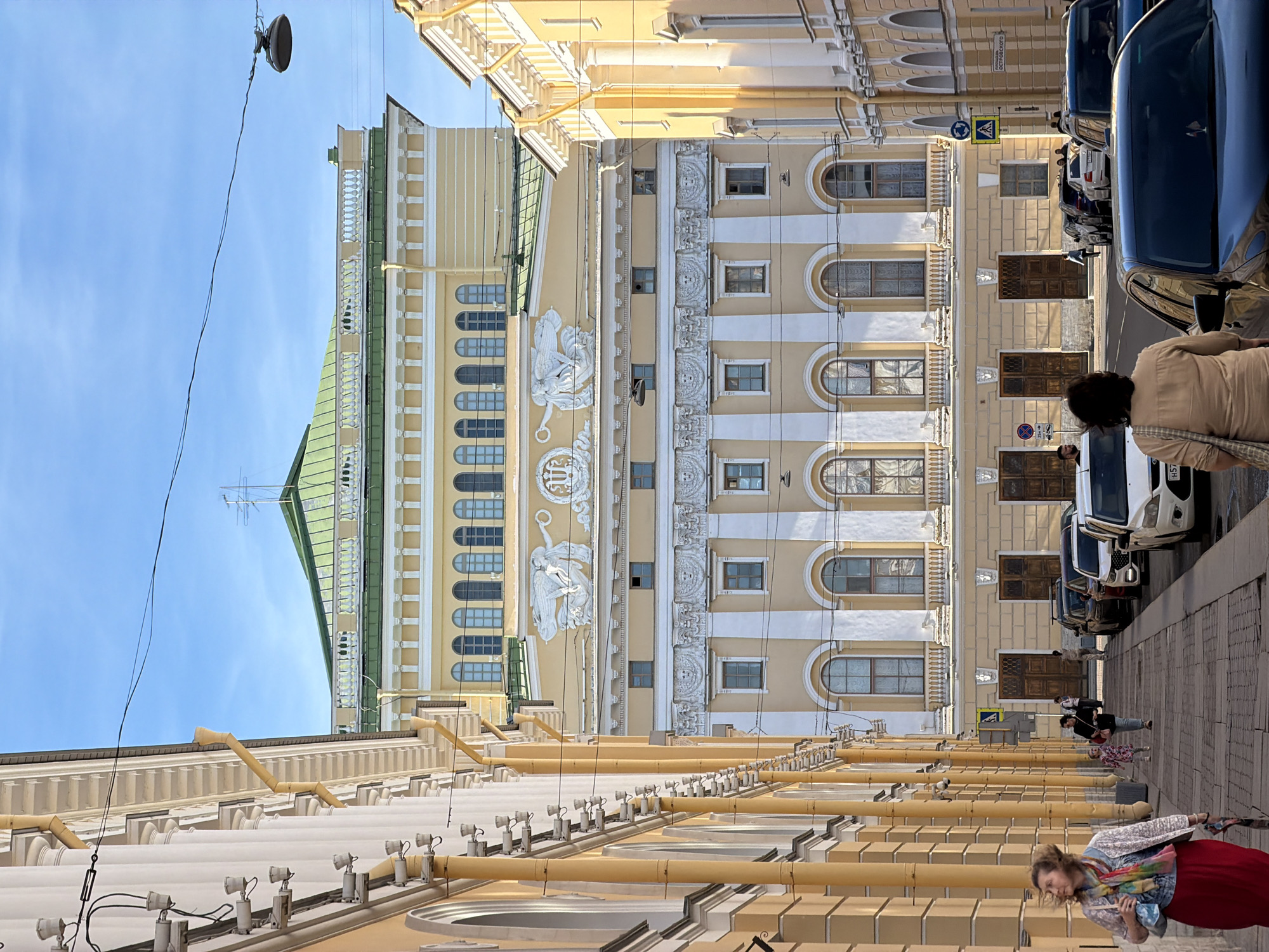 The image depicts a street scene in Saint Petersburg, Russia, looking towards a large, ornate yellow building with a green roof. The building features classical architectural elements, including columns, arched windows, and decorative reliefs. A street lined with parked cars and a pedestrian walkway runs in front of the building. Several people are visible walking on the sidewalk and near the cars. The sky is clear and blue. The architecture suggests a historic district within the city.