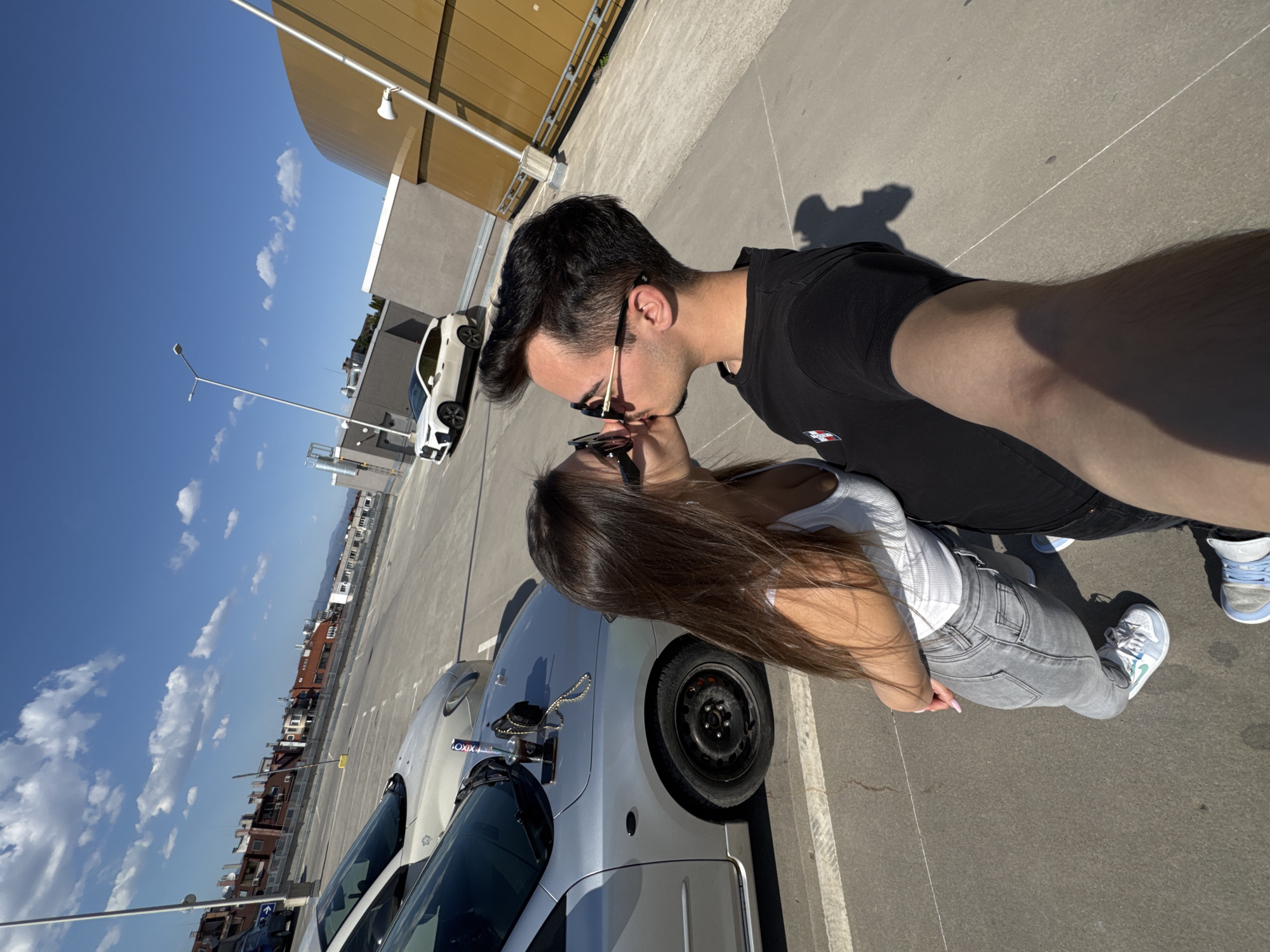 The image depicts a man and a woman kissing in a parking lot. The man has dark hair and is wearing a black t-shirt, sunglasses, and sneakers. The woman has long brown hair and is wearing a white tank top, gray jeans, sunglasses, and sneakers. The parking lot is located in Sofia, Bulgaria. There are cars parked nearby, including a white car in the background and silver cars in the foreground. A tall, modern building and street lights are visible in the background. The sky is blue with scattered clouds, indicating a sunny day.