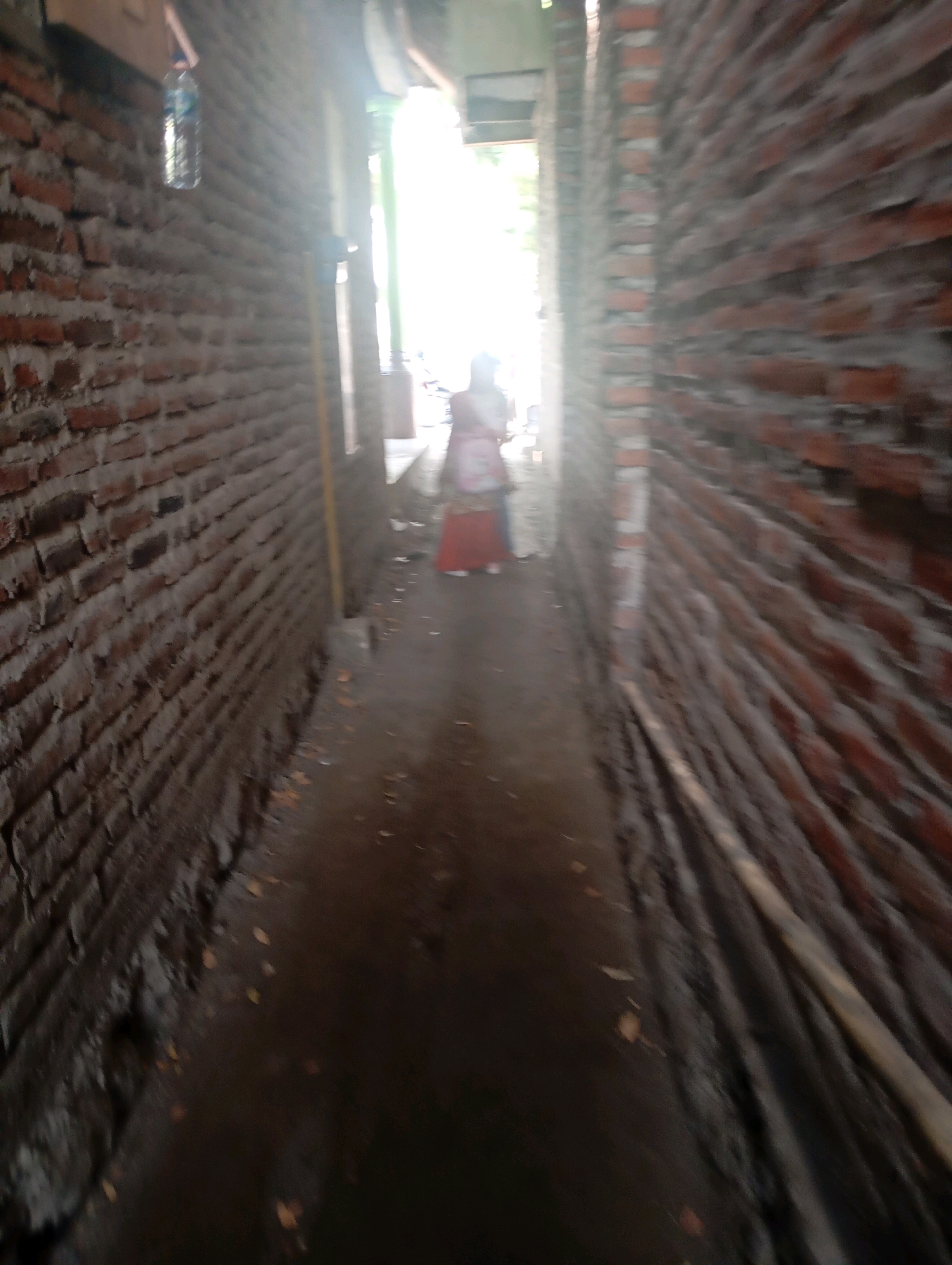 The image shows a narrow outdoor alleyway with brick walls on both sides. The ground is dirt and appears to be wet, with some scattered leaves. In the background, a person is visible, partially obscured by light and motion blur. The person is wearing clothing that appears to be a pink and red garment. The alleyway leads to an area with bright natural light. The overall impression is that the image was taken from a moving perspective, contributing to the motion blur. The location is specified as the city of Ketegan, Indonesia.
