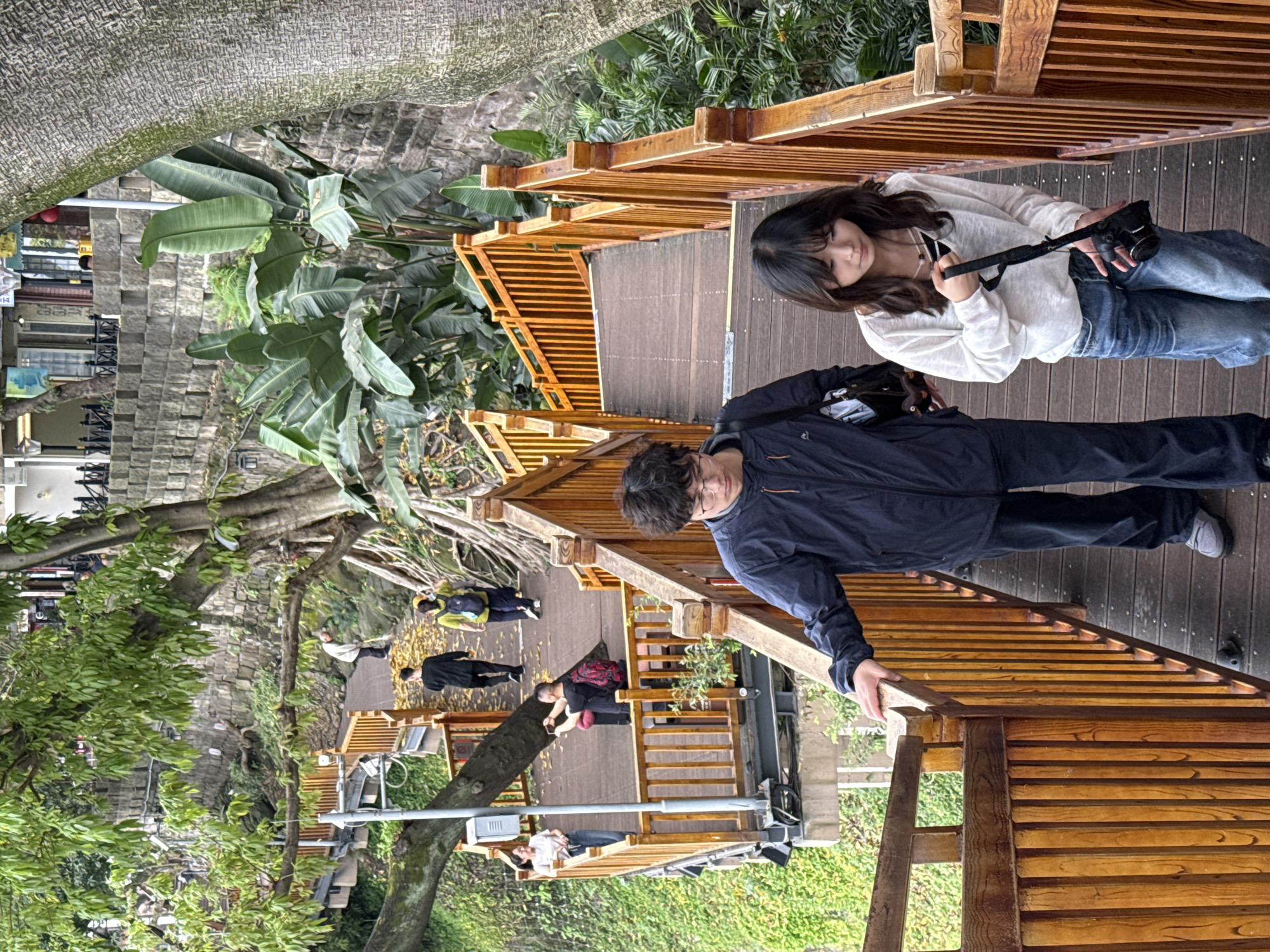 The image depicts a partially obscured outdoor scene in Yuzhong District, Chongqing, China. A wooden boardwalk with railings winds through a lush, green environment. A man in a dark jacket and a woman in a white sweater and jeans are walking along the boardwalk. The woman holds a camera. Other individuals are present along the walkway in the distance. Trees and greenery surround the boardwalk, which is elevated above ground level. A utility pole is visible adjacent to the boardwalk. Buildings can be seen in the background.