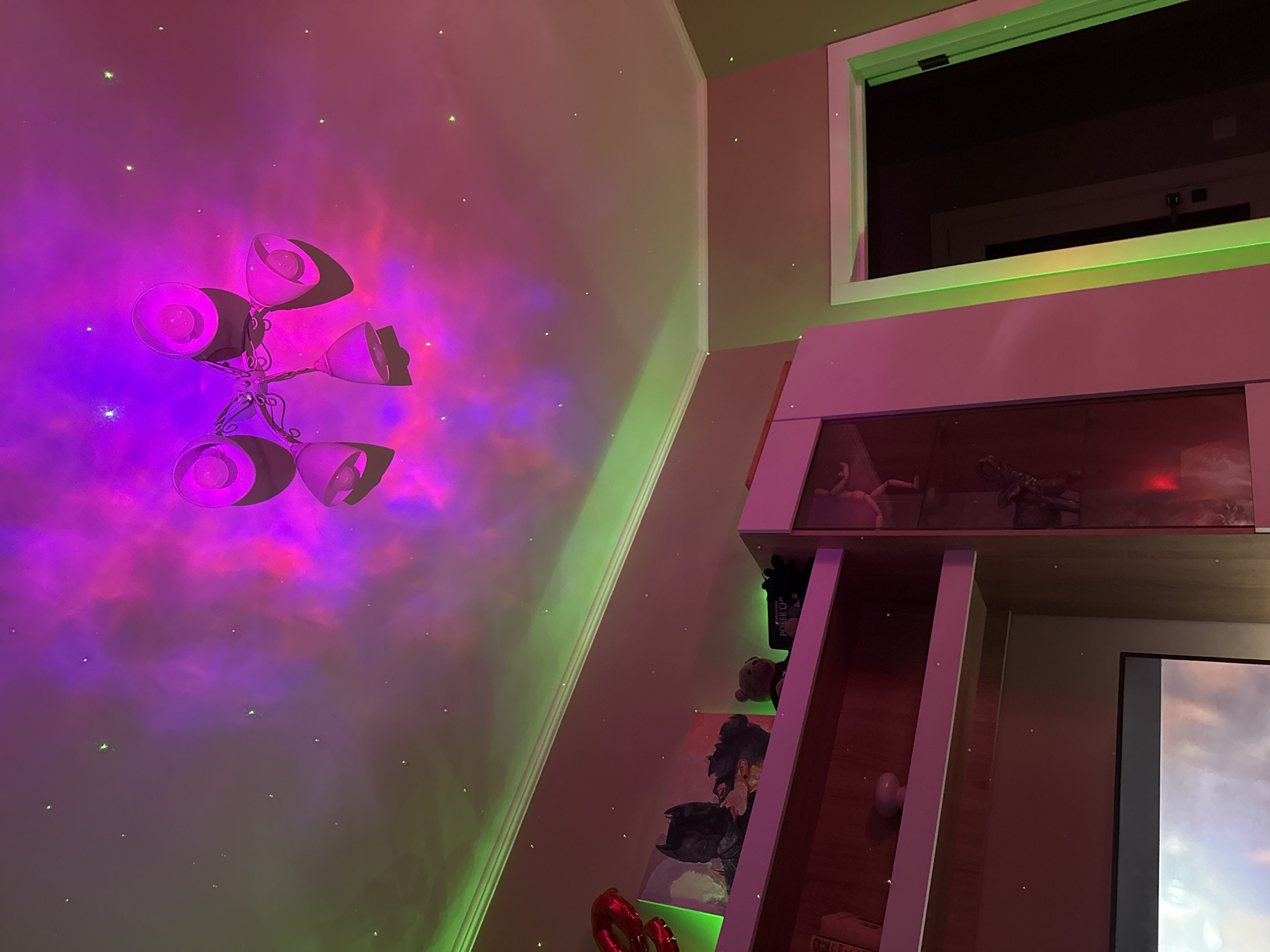 A low-angle indoor shot depicts a room illuminated with neon lighting effects. The left wall is bathed in a purple and pink nebula-like projection interspersed with small white "stars," above a green light strip near the ceiling. A multi-armed chandelier-style light fixture is affixed to this wall, its shades directing light downwards.
To the right, there is a built-in structure with a window above, framed in white with a black interior, reflecting an internal view. The structure features shelves holding items, including a "Poker CP" box and a teddy bear. Below the window, there is a shelf-like enclosure that appears to contain statuettes or figurines. Adjacent to this, there is a partial view of a screen displaying clouds. The lighting in the room casts a pinkish hue over the structure and wall. The location is not possible to determine from the image.