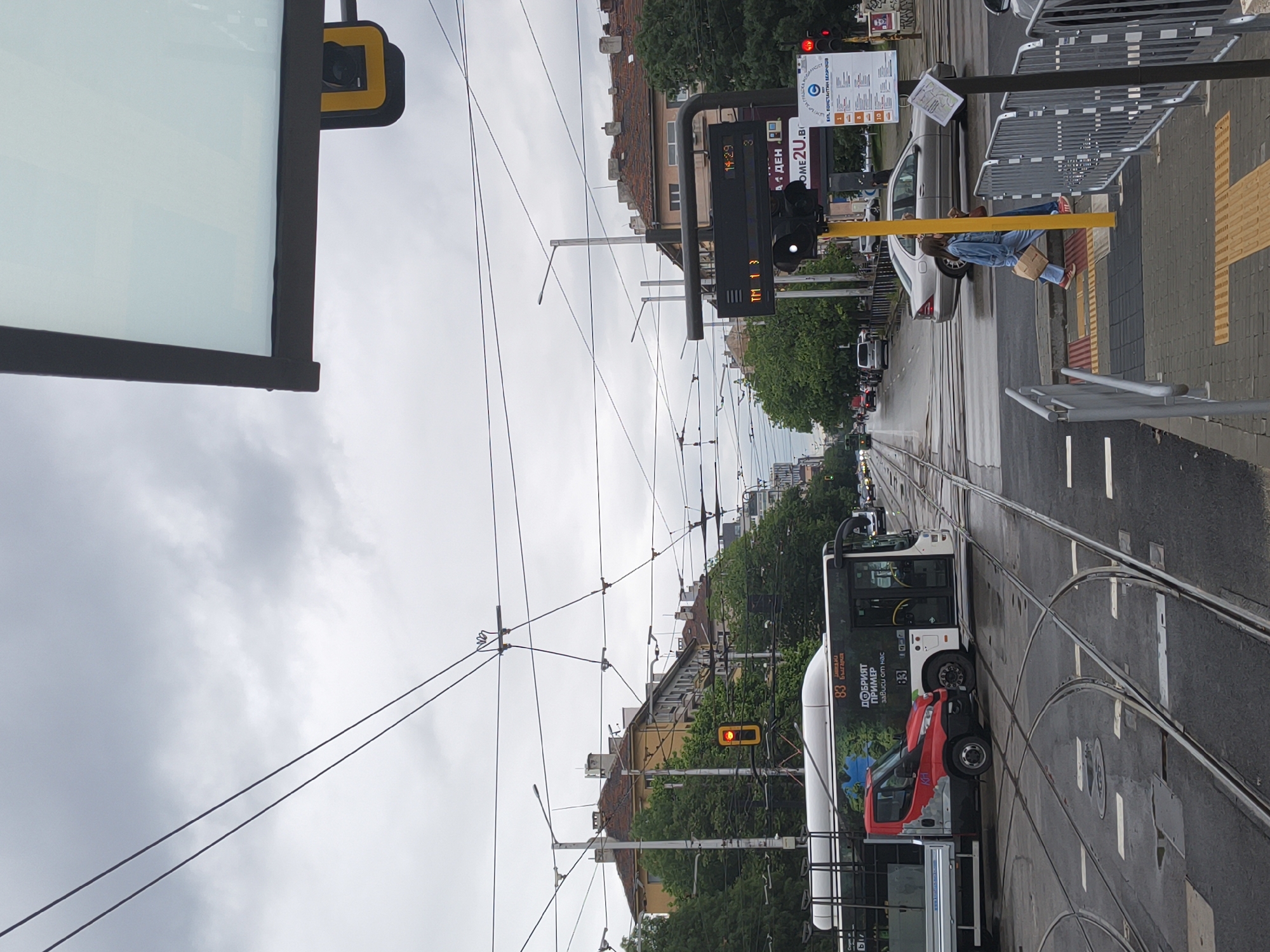 The scene captures an urban landscape in Sofia, Bulgaria, featuring a tram line area during overcast conditions. The tram tracks curve slightly, guiding a tram/bus with an ad displaying "83 БЪЛГАРИЯ" ("83 BULGARIA") and "ДОБРИЯТ ПРИМЕР зависи от нас" ("The GOOD EXAMPLE depends on us"). A red vehicle is parked alongside the tram/bus. A traffic light visible above the tram/bus displays a red signal.
Several buildings with red tile roofs line the street, partially obscured by trees. Overhead power lines create a complex web above the street. A pedestrian sits near a crossing signal with a digital countdown indicating "14:29". The signage next to the lights shows "ул. Константин Величков" ("ul. Konstantin Velichkov"). Barriers protect pedestrians on the sidewalk, where textured pavement is visible. A person wearing a denim jacket, blue jeans, and red shoes is sitting against a traffic light, holding a brown square container.
