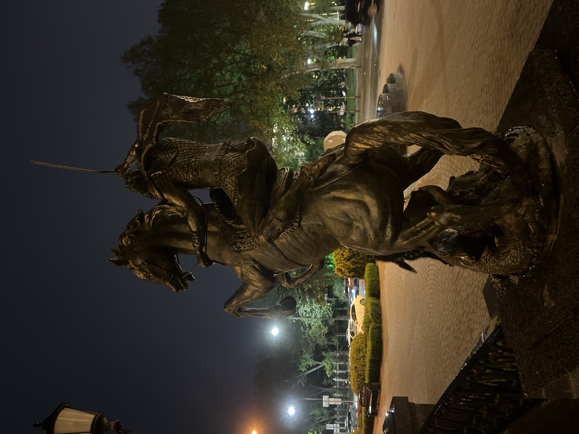 The image shows a statue of a warrior on horseback, with the warrior wielding a sword. The statue is made of dark metal and is set against a backdrop of trees and streetlights. In the background, there is a cobblestone walkway. The lighting suggests it is nighttime. The image shows the statue of Saint George slaying the dragon, located in the city of Krasnodar, Russia.