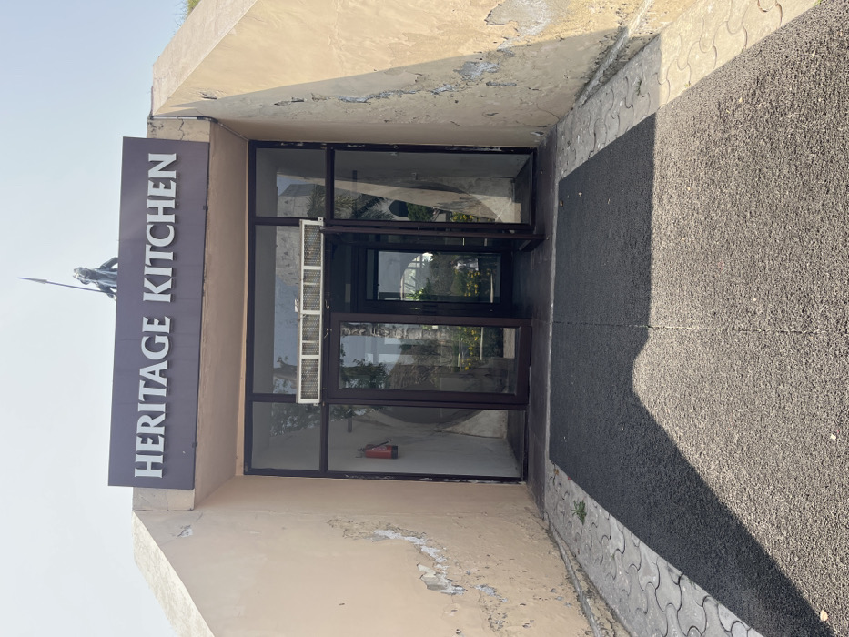 The image shows the entrance to a restaurant called "Heritage Kitchen" in Sahibzada Ajit Singh Nagar, India. The entrance is made of large, glass double doors set in a brown frame. The doors are open. There are large, rectangular windows on either side of the doors. Above the doors is a brown sign with white lettering that reads "Heritage Kitchen". The sign is mounted to a concrete awning. The walls of the building are a pale yellow and show signs of weathering. There is a black mat in front of the entrance. The ground surrounding the entrance is made of cement. There is a red fire extinguisher visible on the left wall. There is a small, brown sculpture on the roof above the sign. The building is located in Sahibzada Ajit Singh Nagar, India, at the coordinates 30.703 latitude, 76.668 longitude.