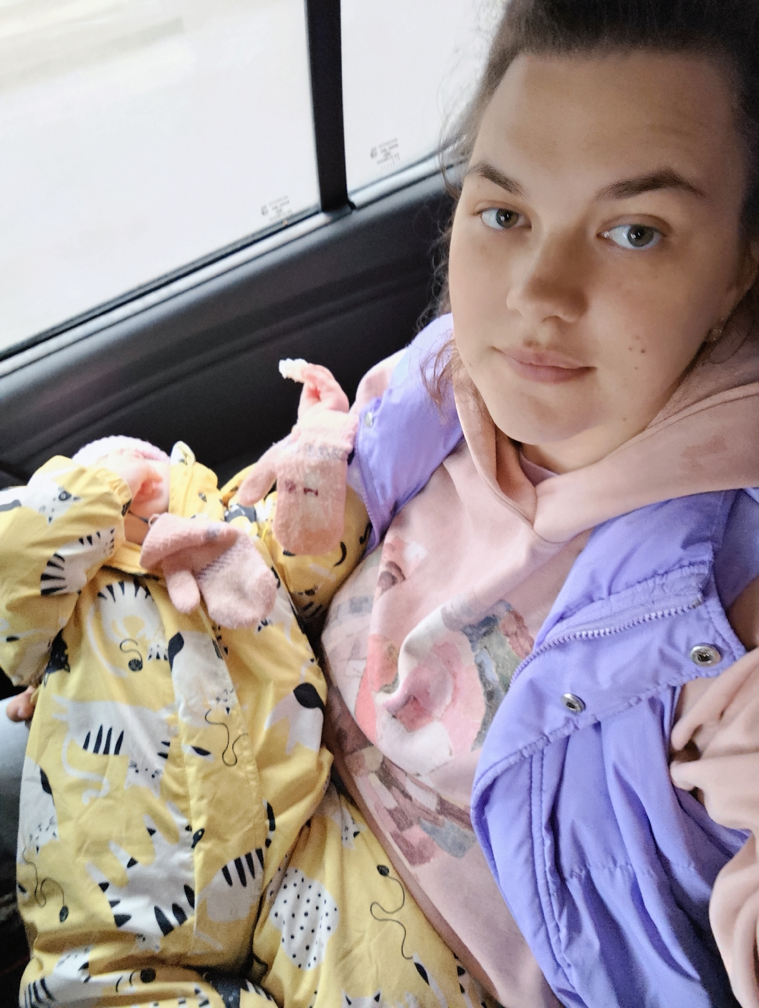 A young woman is seated in the back of a car, presumably in transit within Chaikovsky, Russia. She is dressed in a pink hooded sweatshirt and a light purple puffy vest. To her left is a child's yellow snowsuit featuring a black and white tiger pattern, along with pink mittens. The car's interior is dark, with a window visible in the background. The woman is looking directly at the camera with a neutral expression.